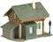 Mały model wiejskiego domu z zielonym dachem, kominem i dobudówką. Obok czarna latarnia uliczna.