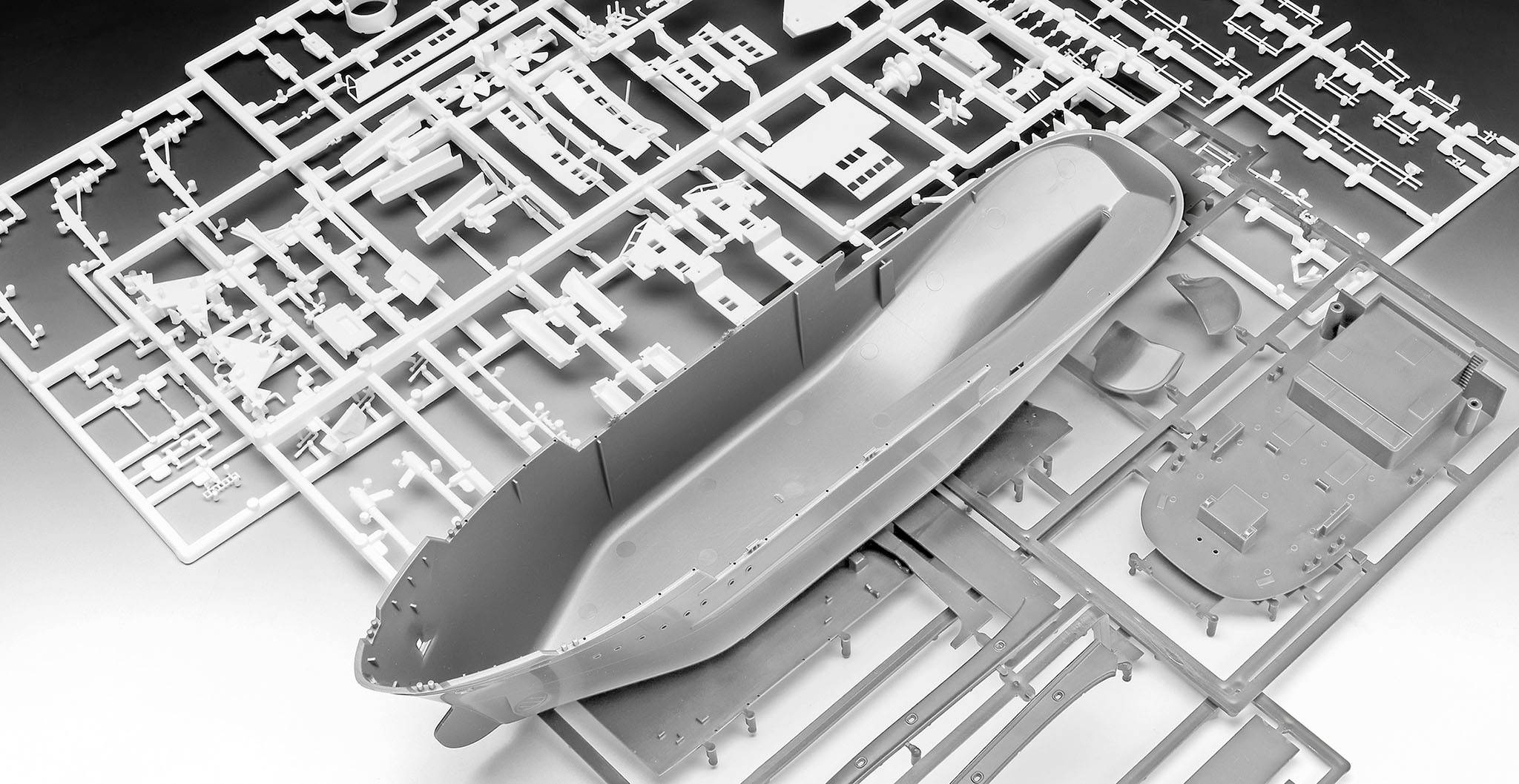 Szary model statku w częściach leży na płycie montażowej. Widoczny jest kadłub i inne komponenty, gotowe do złożenia.