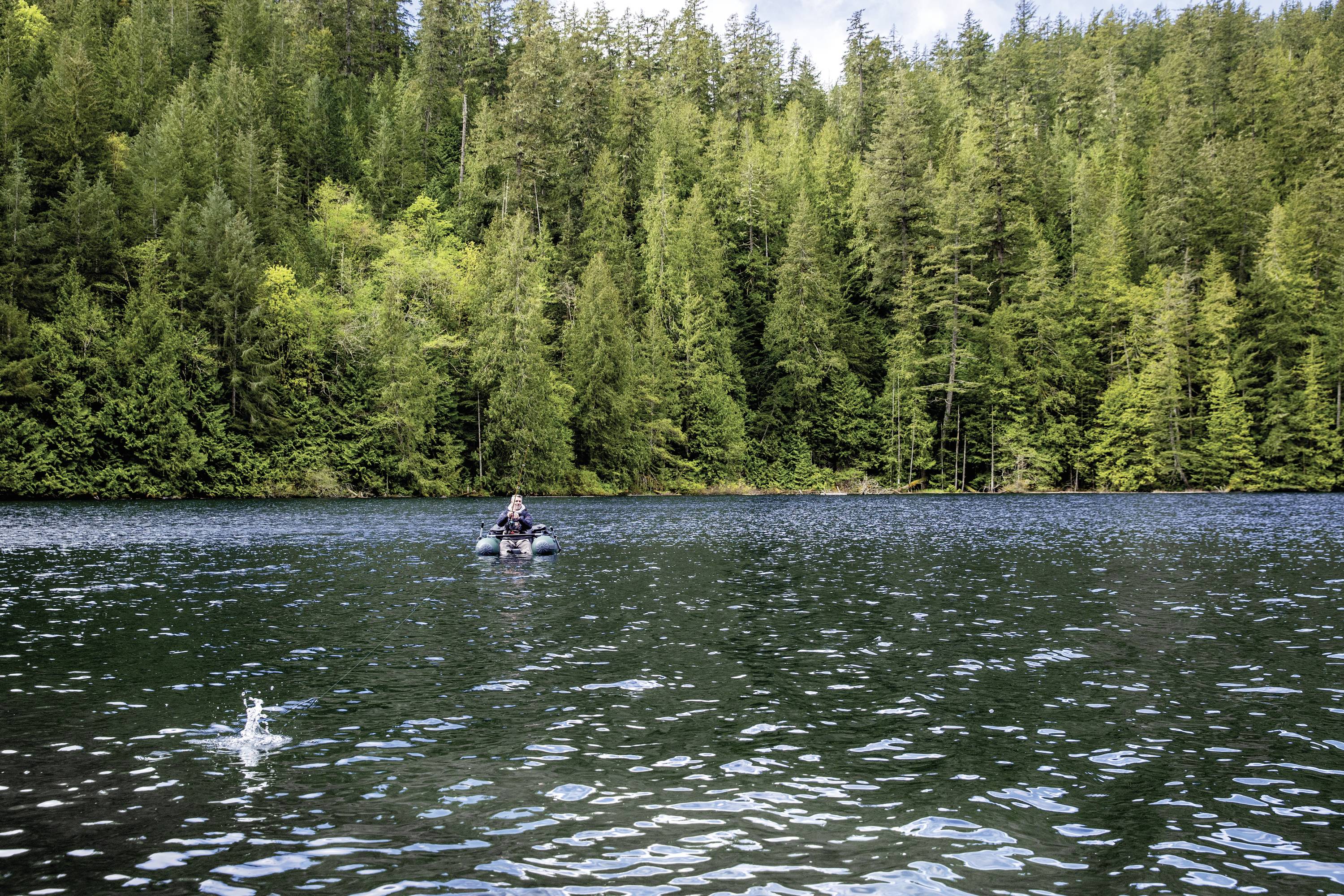 Osoba wędkująca w łodzi na jeziorze, otoczona gęstymi, zielonymi drzewami. Ryba skacze z wody na pierwszym planie.