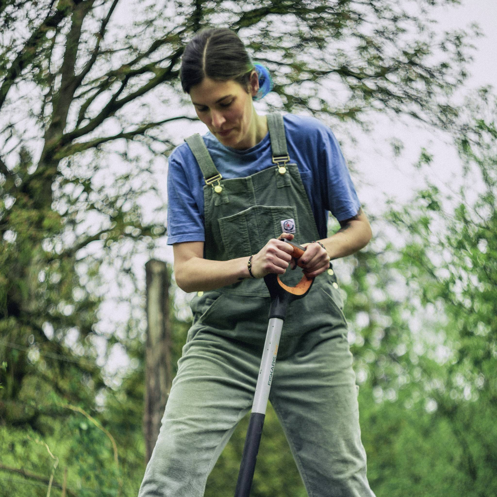 Osoba pracuje w ogrodzie, ubrana jest w niebieskie ubranie i trzyma łopatę. W tle widoczne są drzewa.