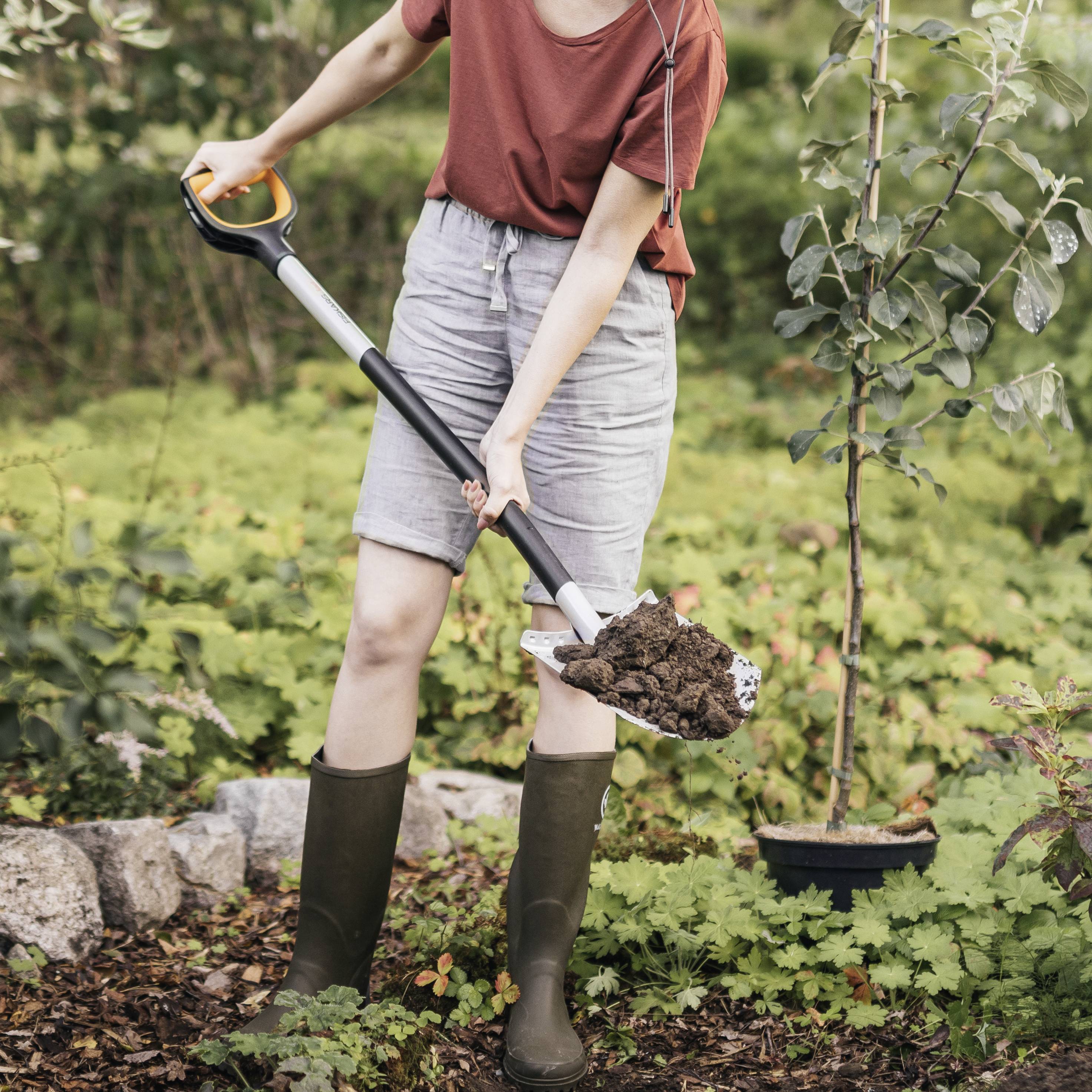 Osoba kopie ziemię w ogrodzie, nosi buty i trzyma łopatę. Otoczona roślinami i kamiennym obrzeżem.