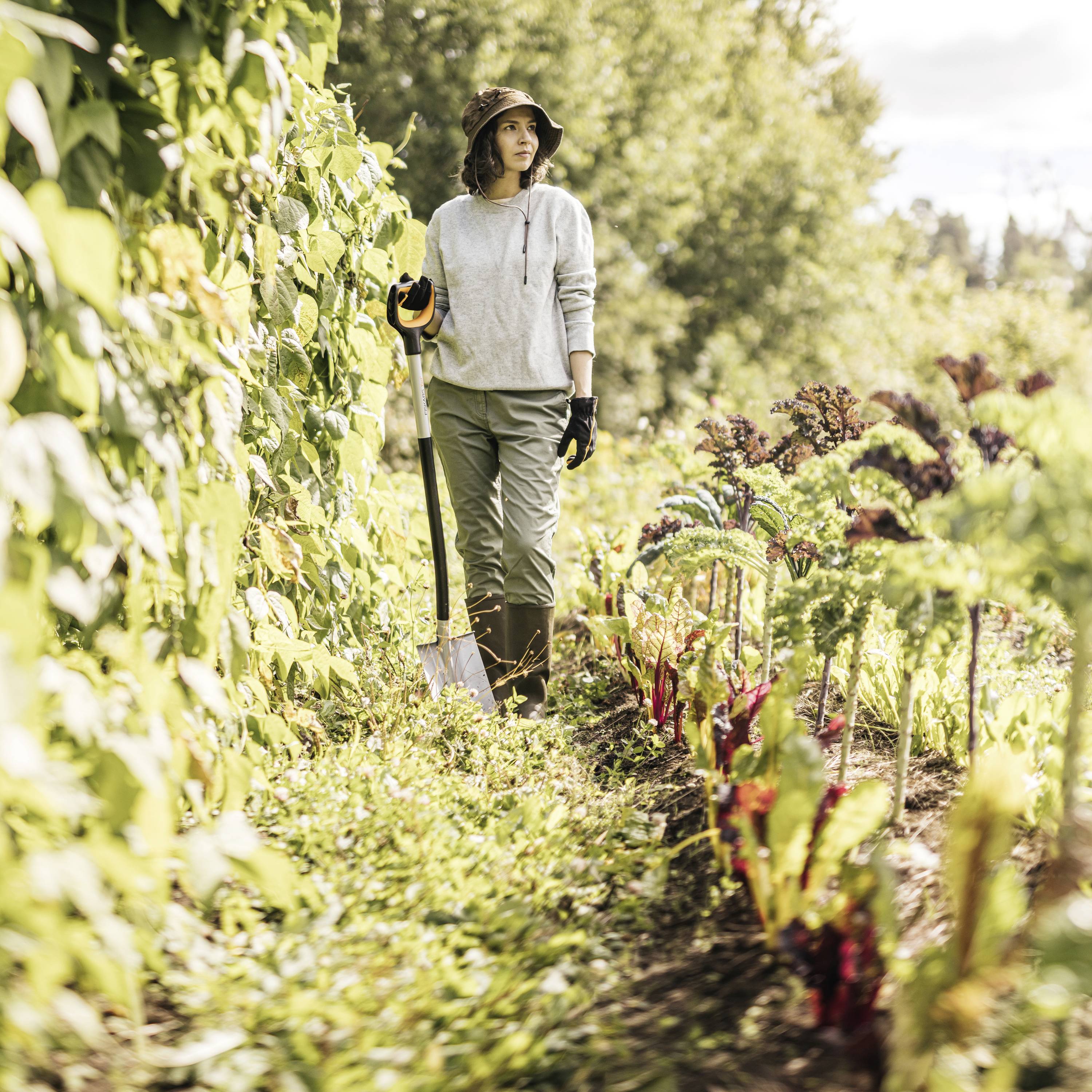 Osoba w kapeluszu przeciwsłonecznym stoi w ogrodzie warzywnym, trzymając łopatę. Kolorowe rośliny i drzewa rozświetlają otoczenie.