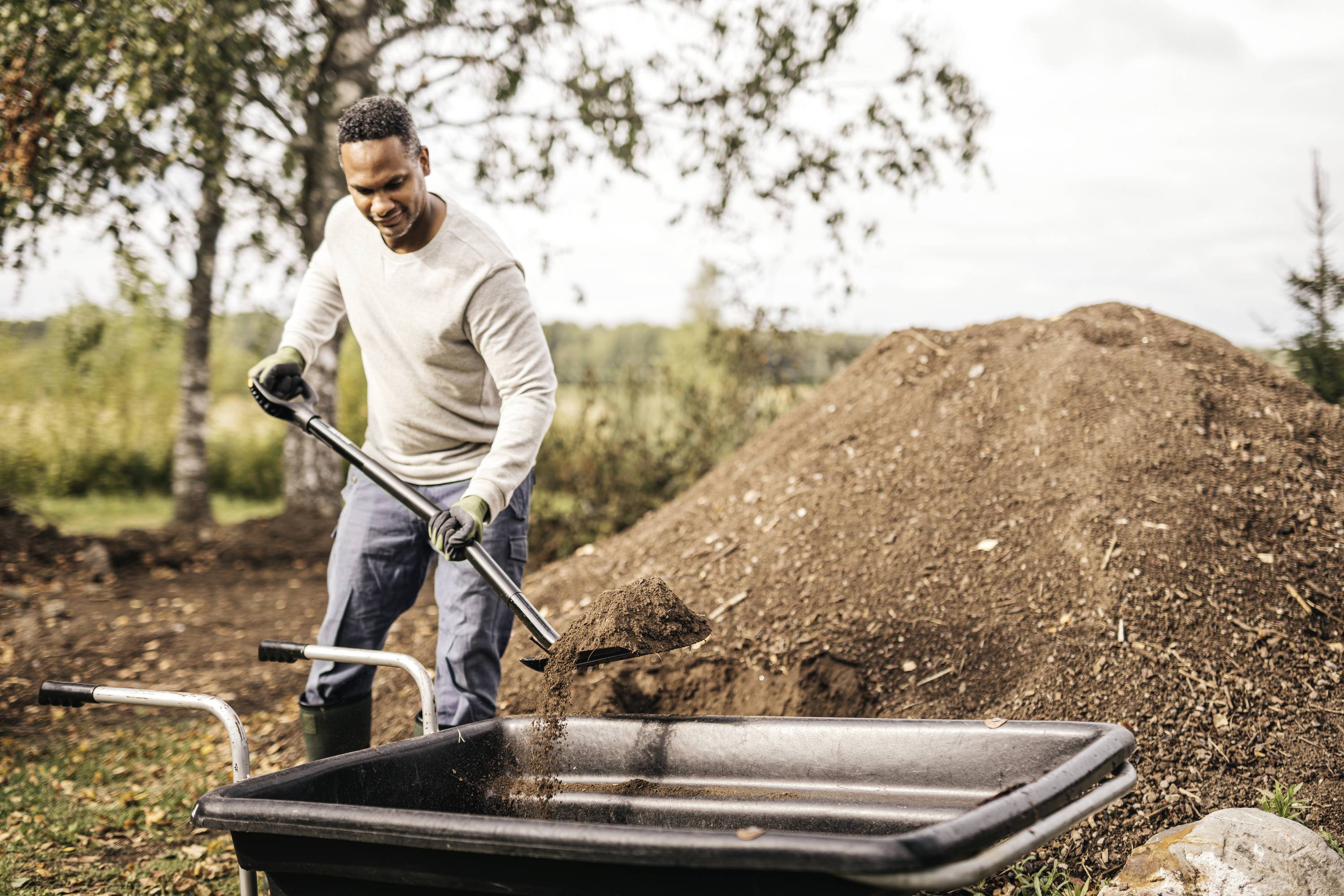 Mężczyzna ładuje ziemię do taczek na zewnątrz, przed dużą hałdą ziemi. W tle widoczne są drzewa.