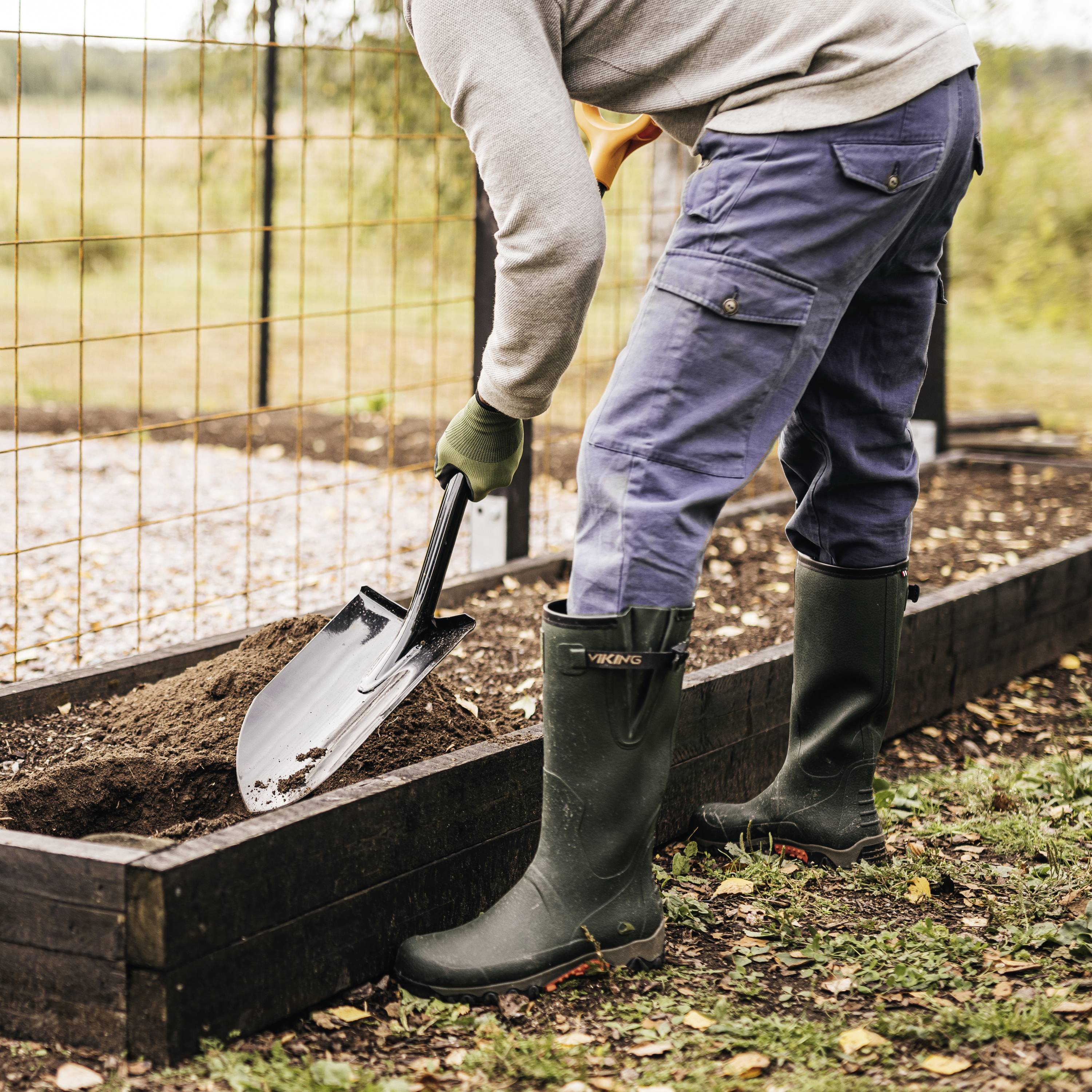 Osoba kopie ziemię łopatą w grządce ogrodowej. Ma na sobie gumowce i stoi przy metalowym ogrodzeniu.