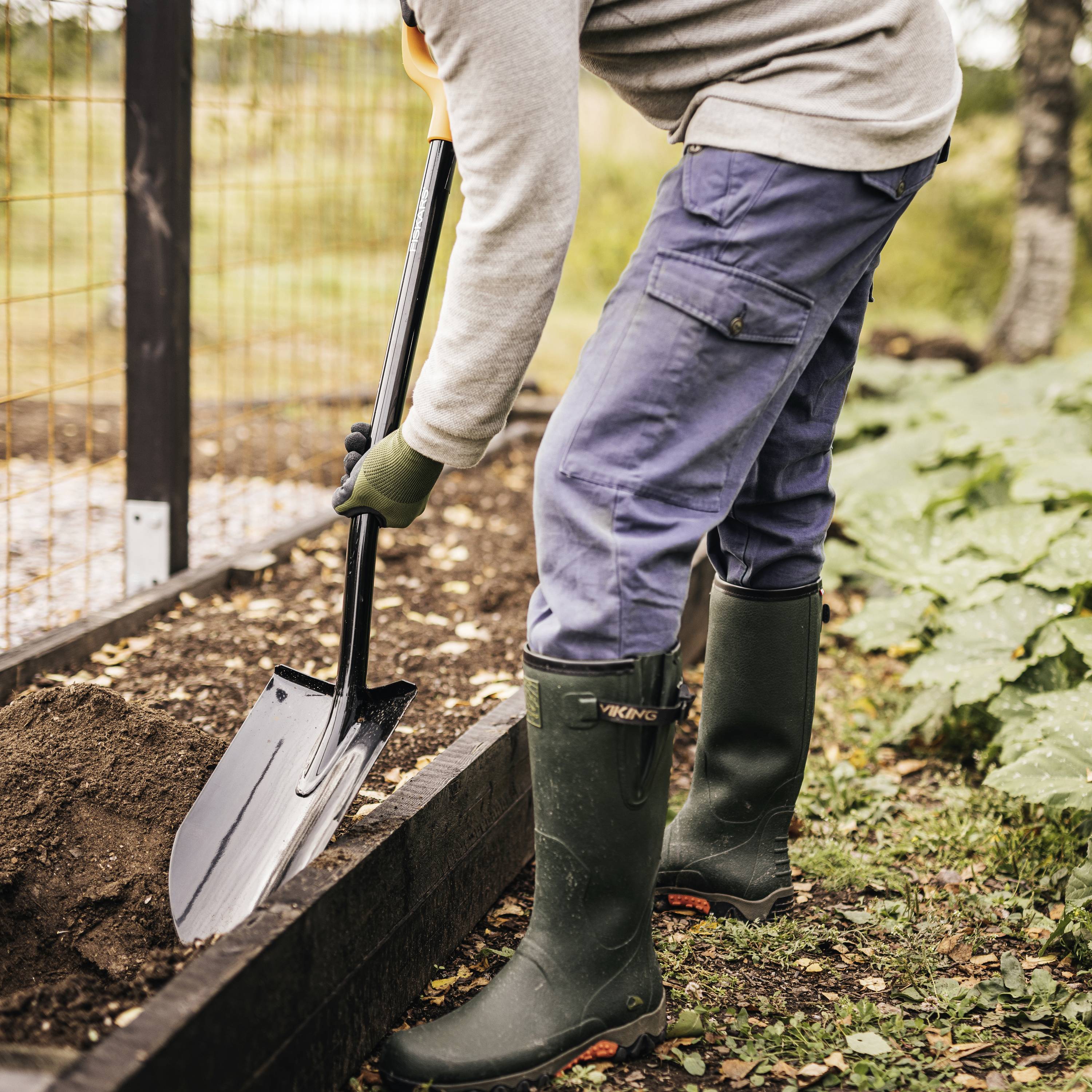 Osoba kopie ziemię łopatą w ogrodzie, ubrana w kalosze i spodnie robocze. W tle drewniany płot i rośliny.
