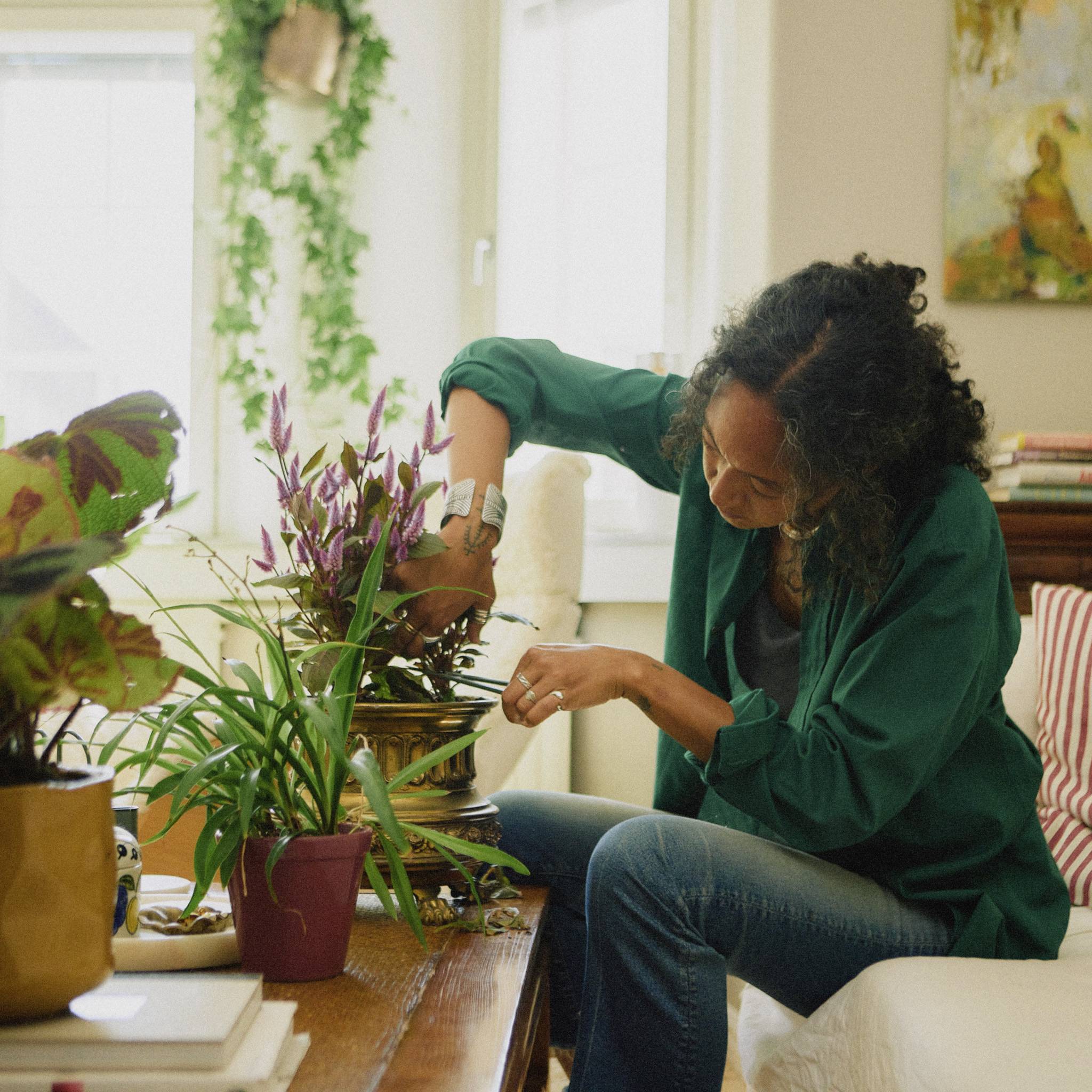 Osoba siedzi przy stole i pielęgnuje rośliny w pomieszczeniu pełnym zieleni, tworząc spokojną i relaksacyjną atmosferę.