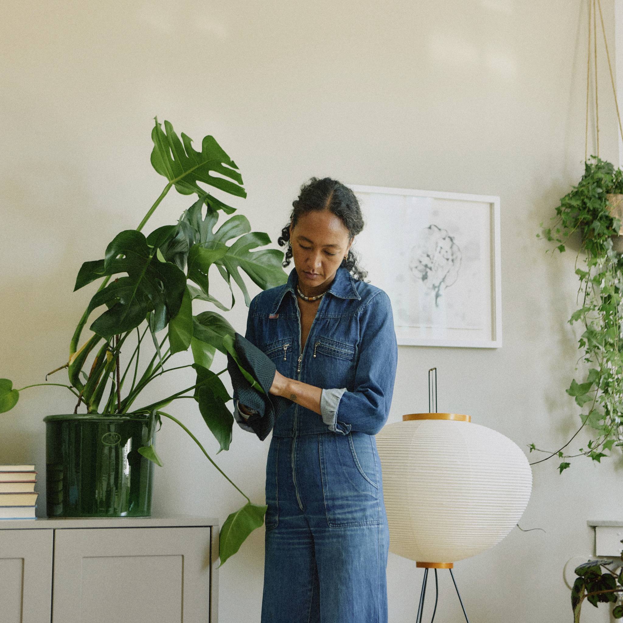 Osoba w jeansowym stroju stoi obok dużej rośliny i okrągłej lampy, na jasnym tle ozdobonym dziełami sztuki.