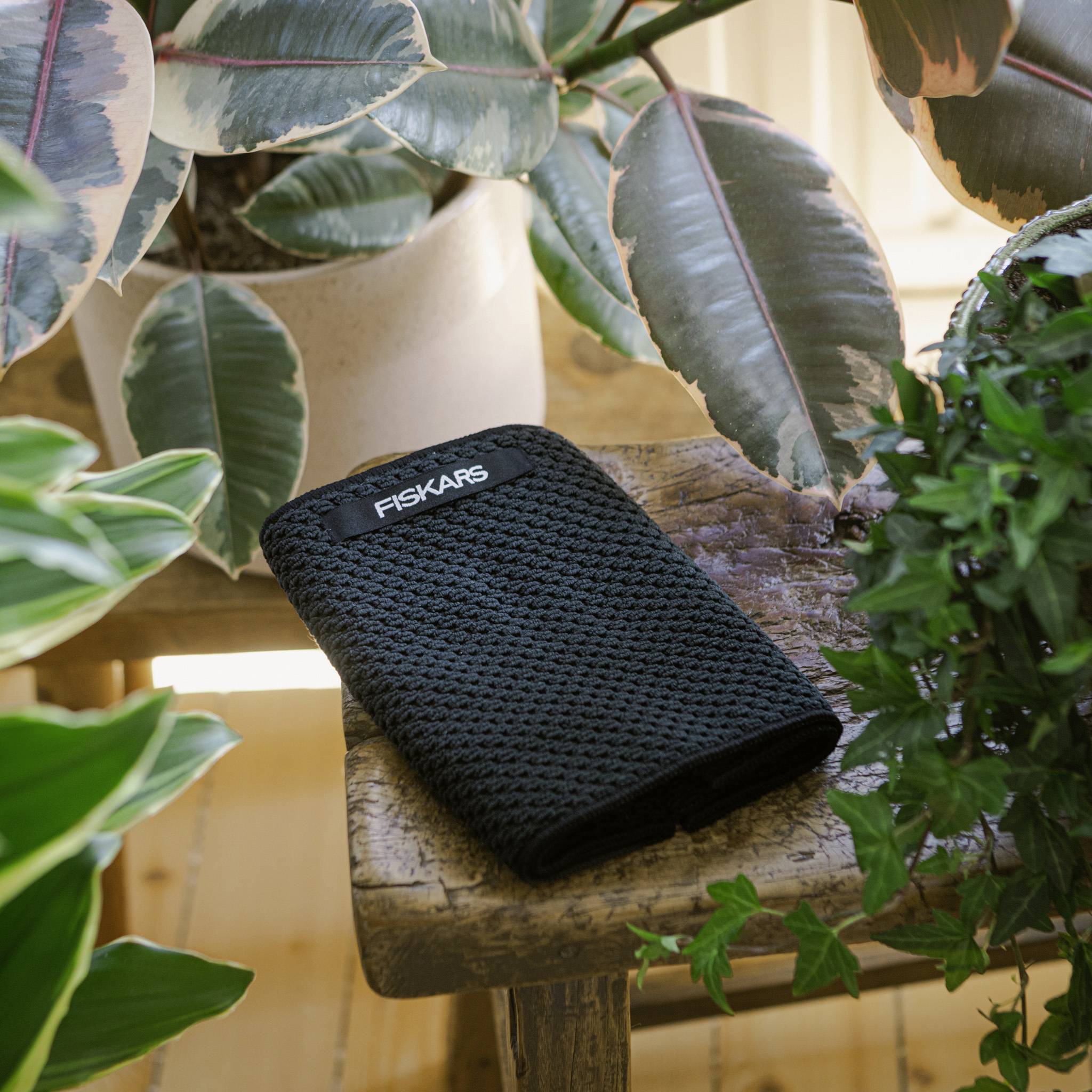 Czarne narzędzie ogrodnicze z logo 'Fiskars' leżące na drewnianym stole, otoczone różnymi roślinami tropikalnymi w doniczkach.