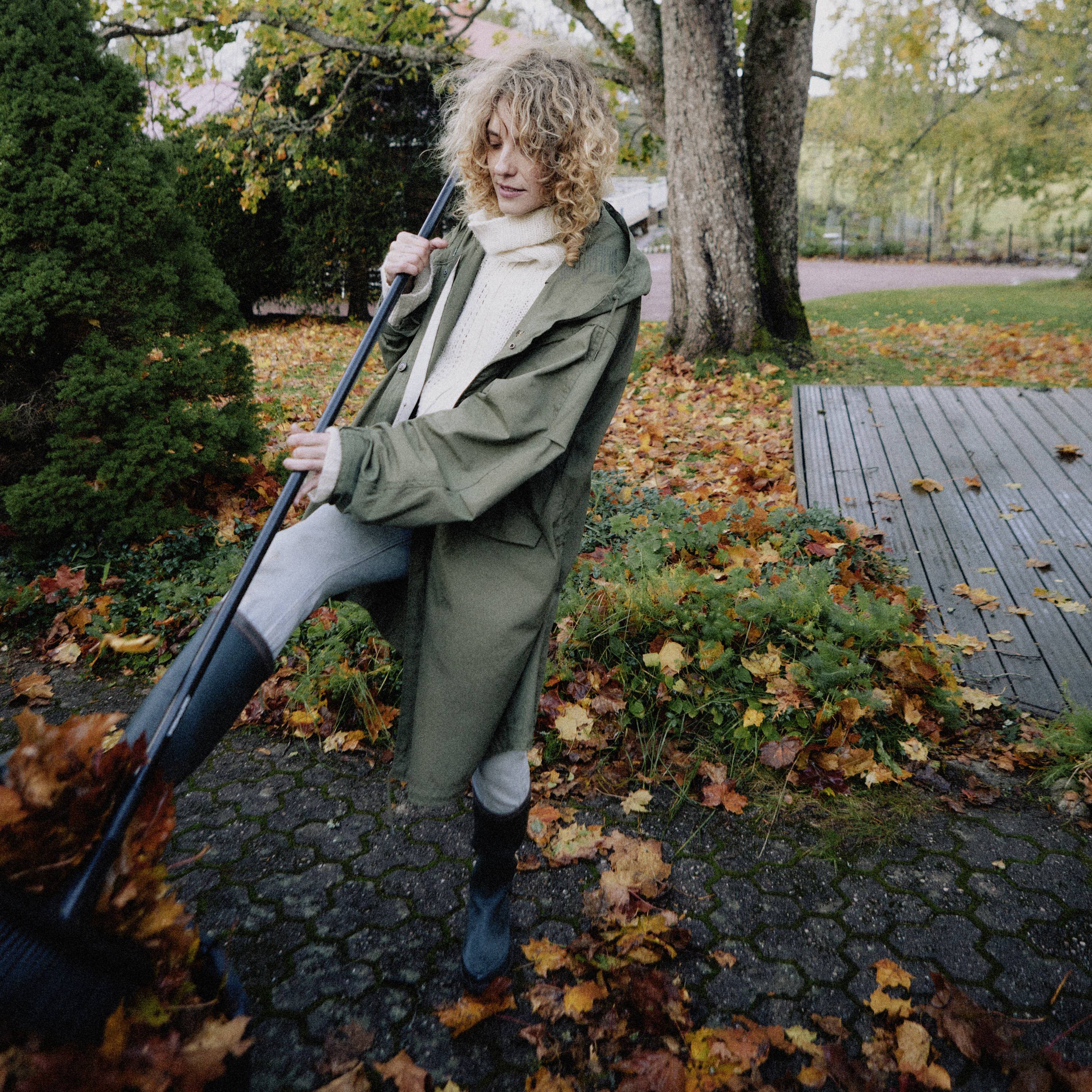 Osoba w zielonym płaszczu zamiatająca liście na chodniku jesienią. W tle widoczne są drzewa pokryte kolorowymi liśćmi.