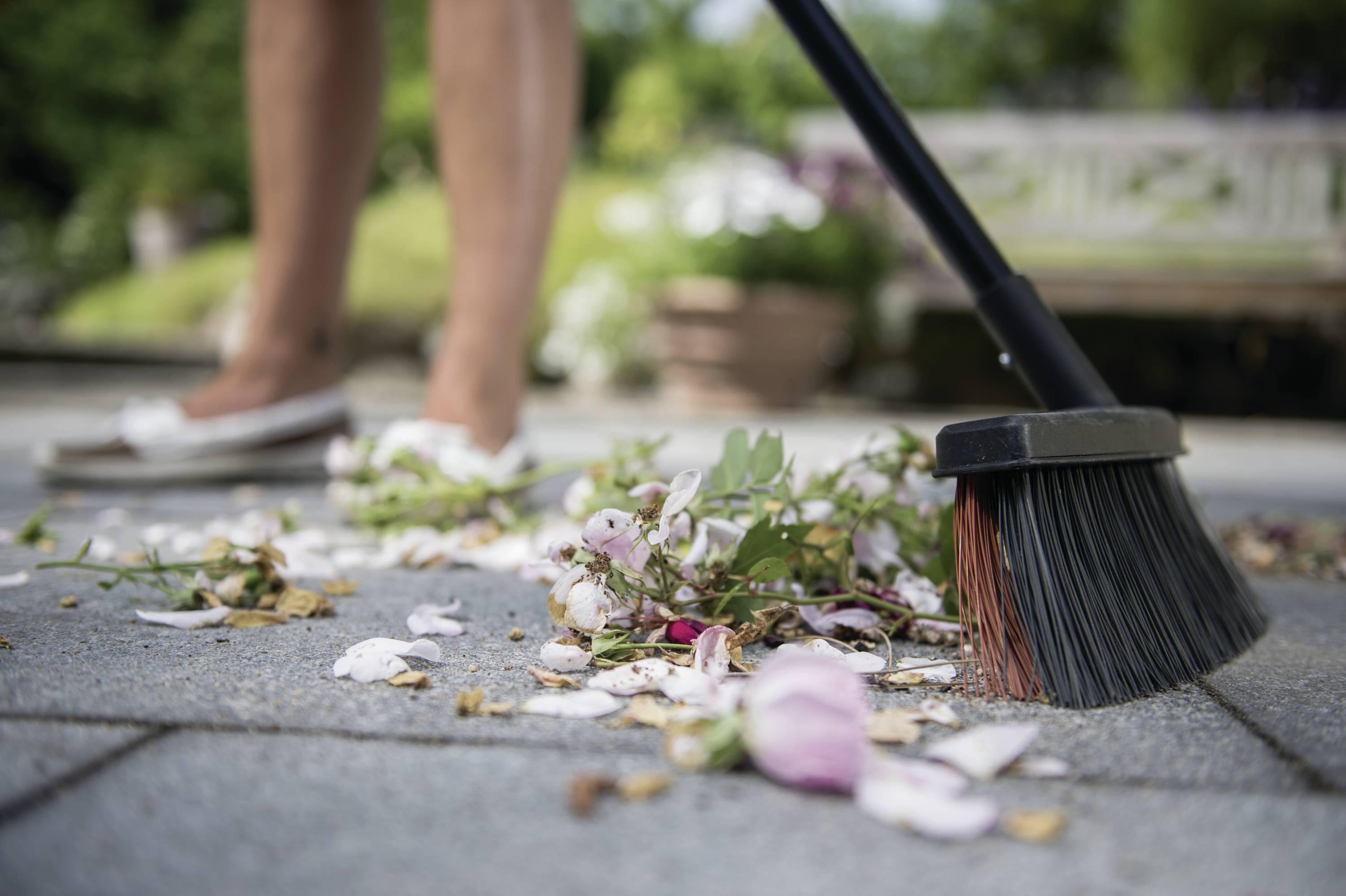 Osoba zamiata płatki kwiatów i liście z tarasu. W tle widać rozmyte rośliny ogrodowe i drewnianą ławkę.