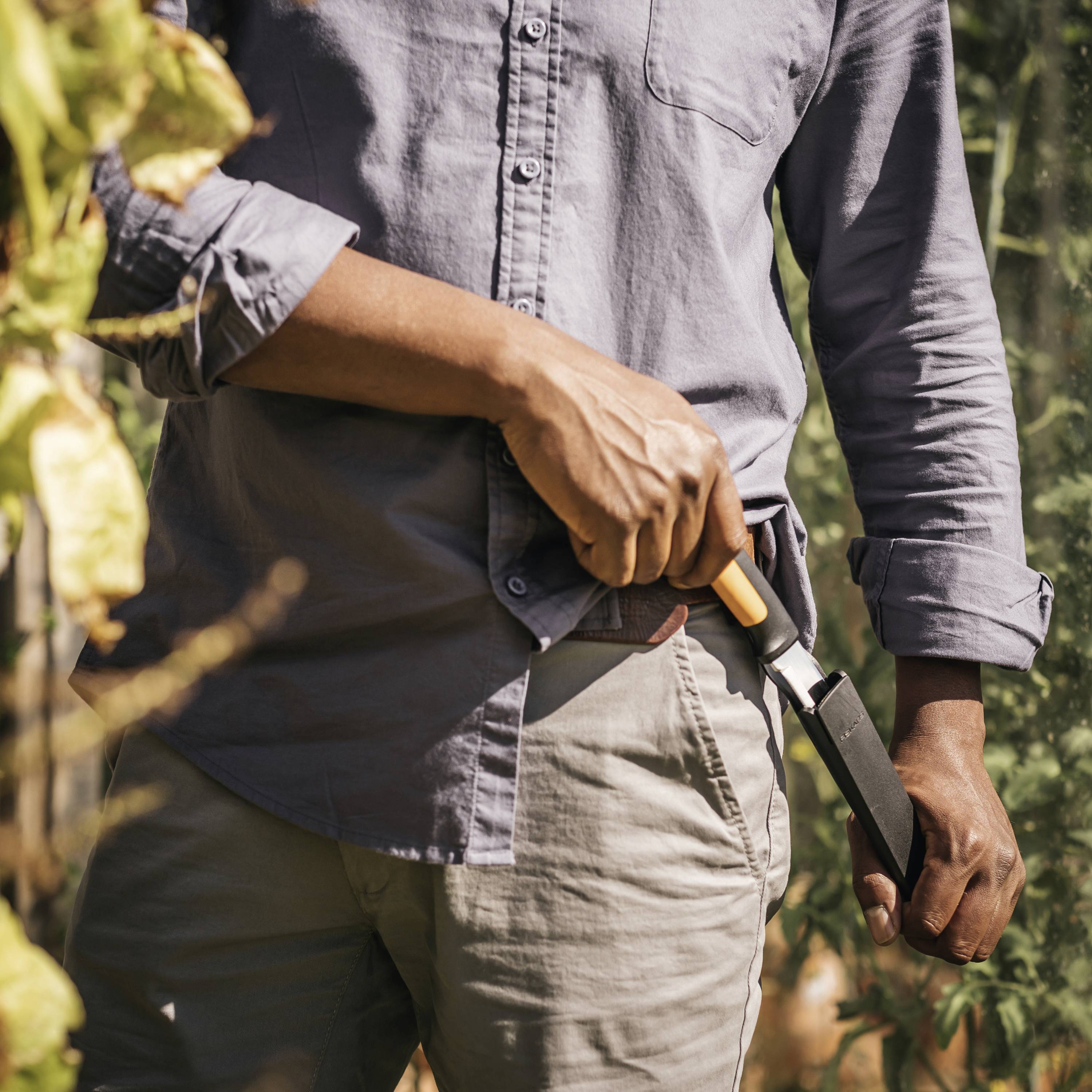 Osoba w koszuli przycina rośliny w słońcu, trzymając nóż w prawej ręce. Chwila pracy w ogrodzie jesienią.