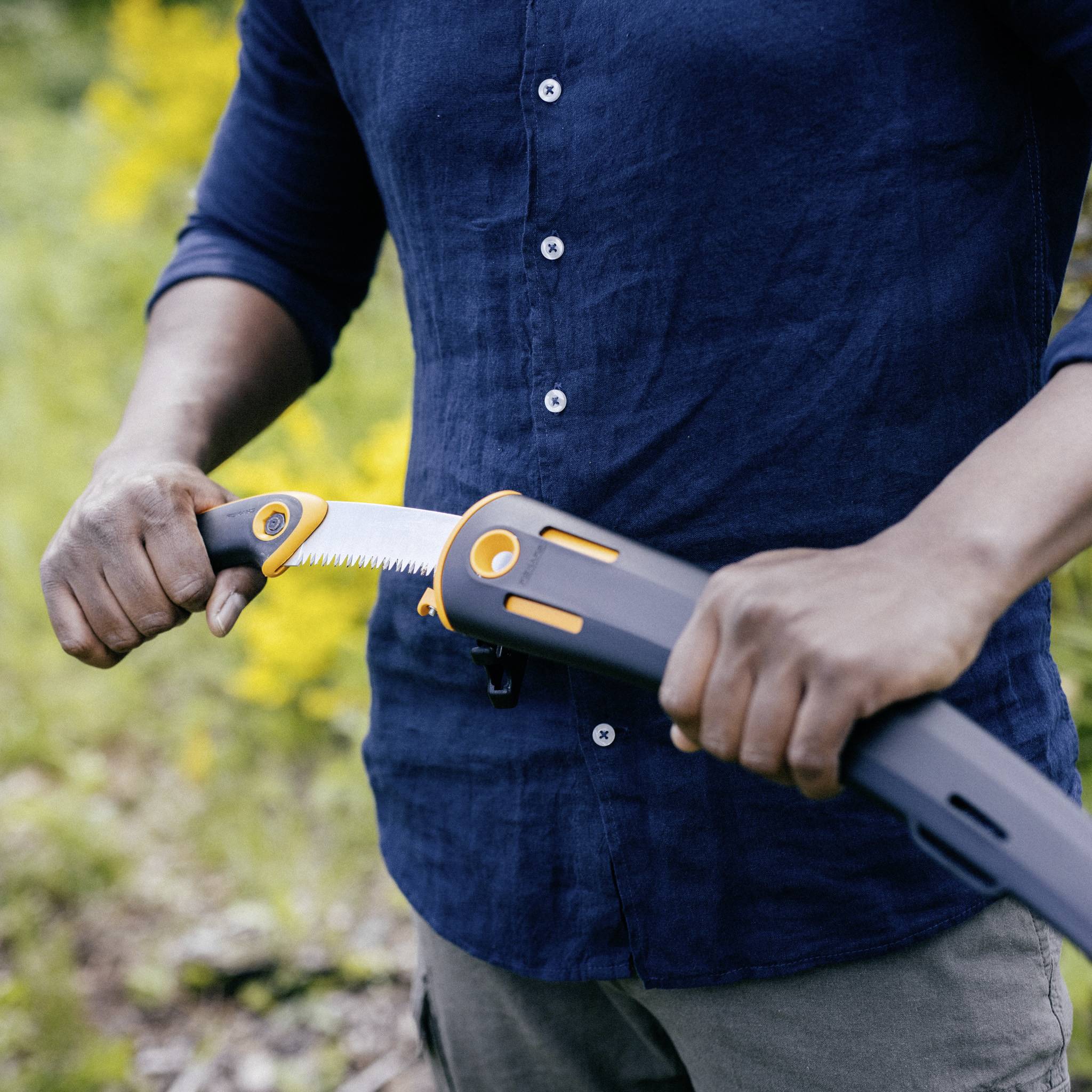 Osoba przecina gałąź składaną piłą ręczną w kolorze pomarańczowo-szarym na wolnym powietrzu. W tle widać rozmyte zielone roślinności.