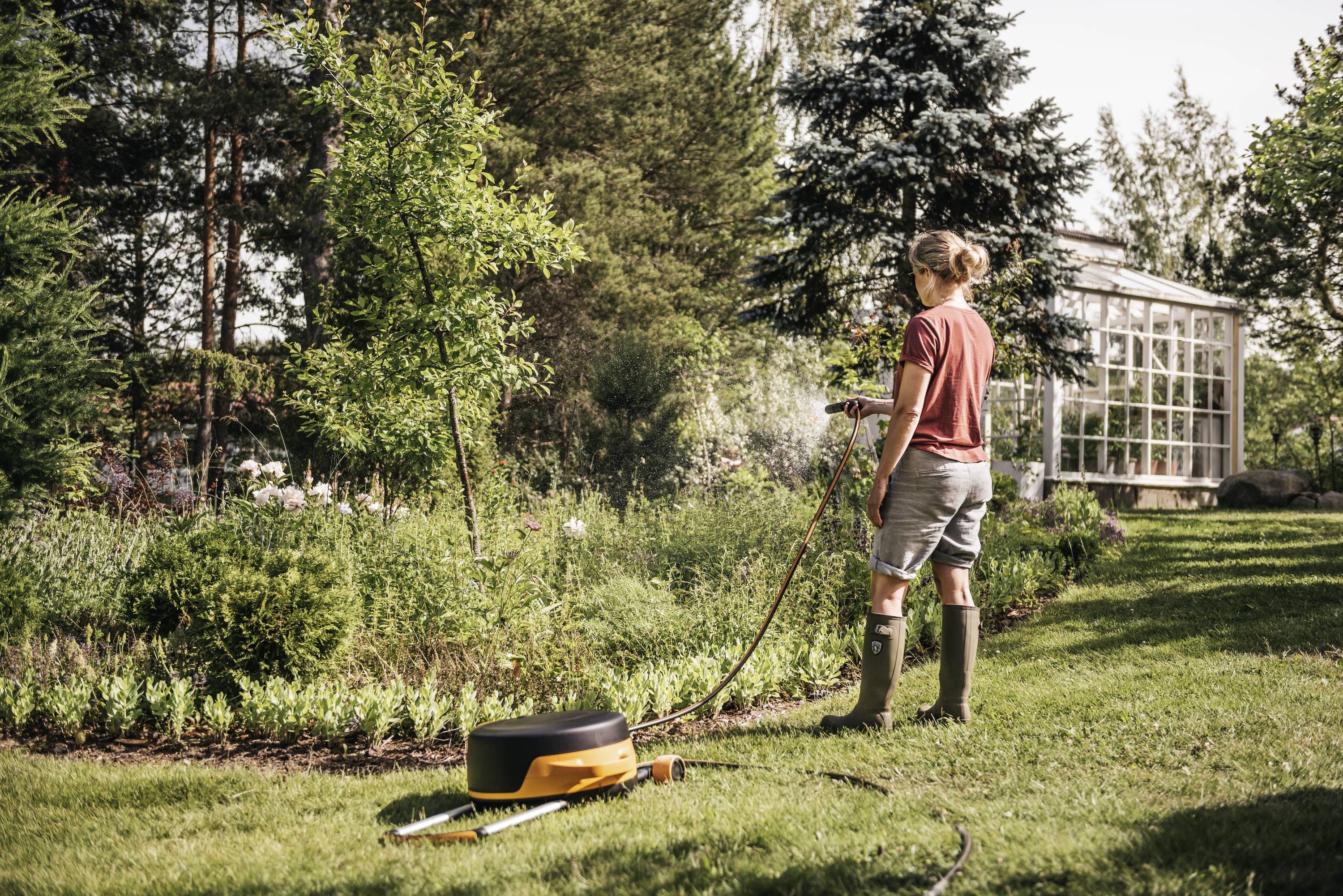 Osoba podlewa rośliny w zielonym ogrodzie obok szklarni. W tle widać wysokie drzewa.