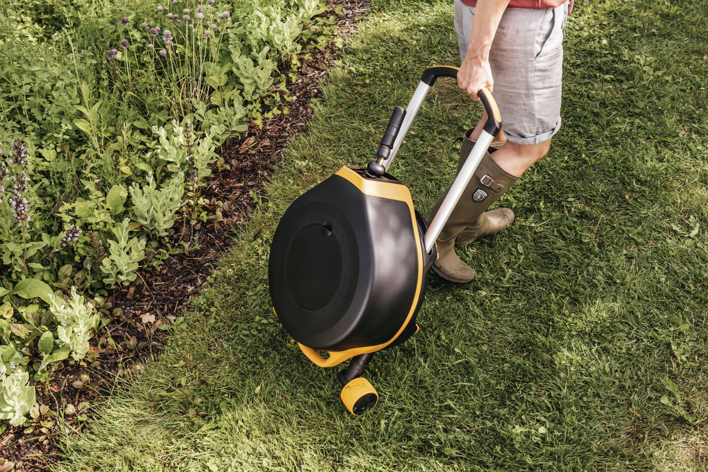 Osoba kosi trawę żółtą kosiarką ręczną na łące obok grządki ogrodowej.