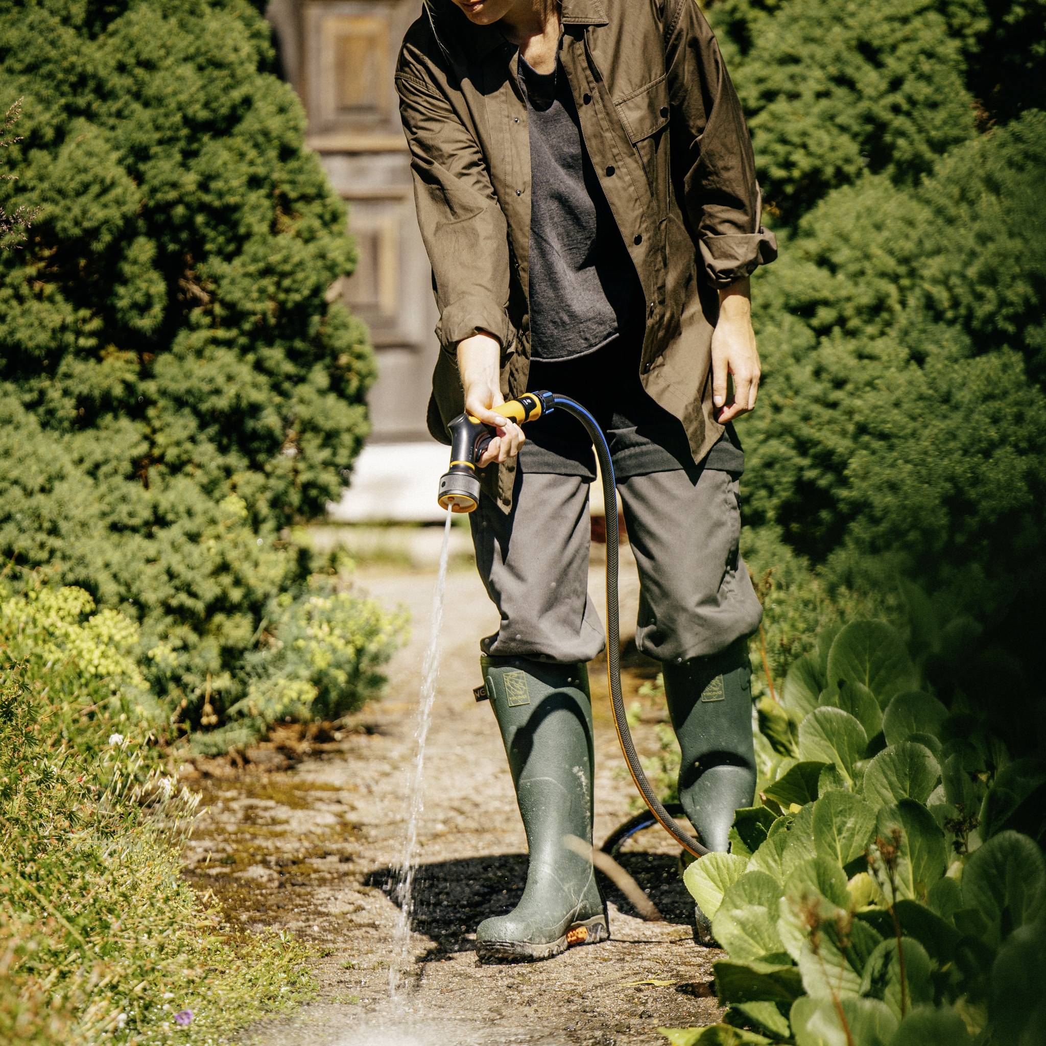 Osoba podlewa ścieżkę ogrodową za pomocą węża. Ma na sobie gumowce i jest otoczona zielonymi krzewami i roślinami.