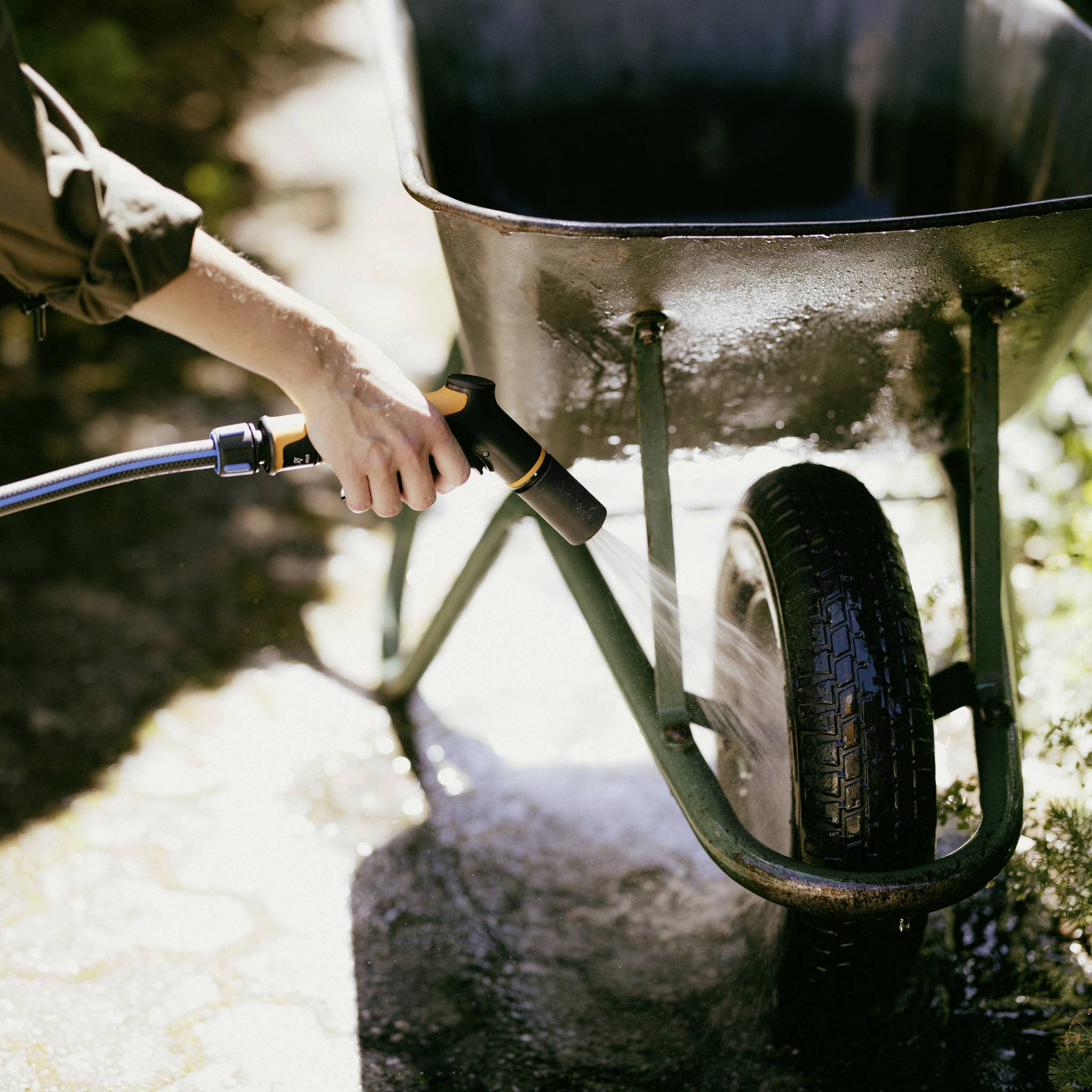 Osoba polewa wodą z węża koło taczek. Scena rozgrywa się na słonecznym podwórku.