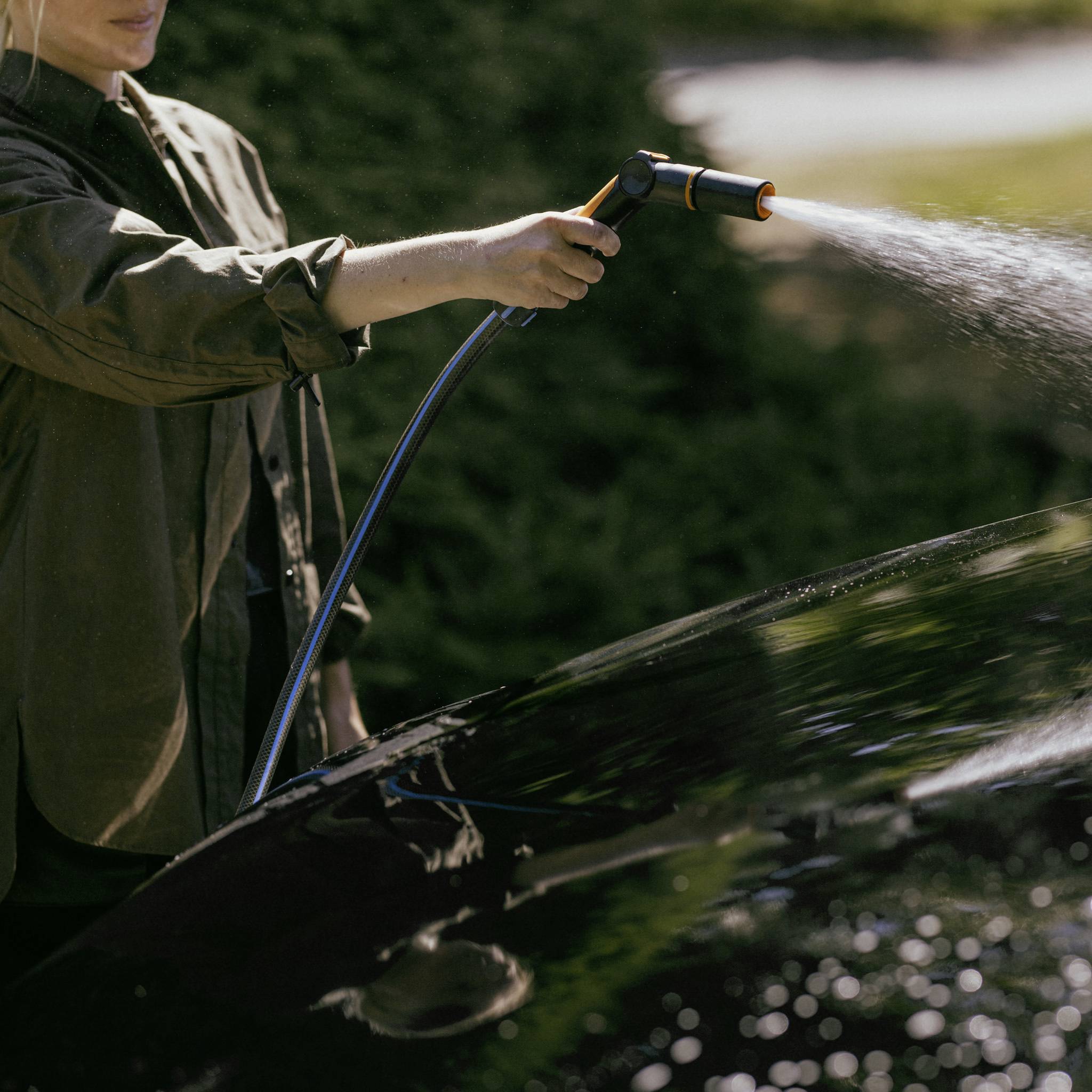 Osoba myje samochód wężem ogrodowym. W tle widać nieostre krzewy.