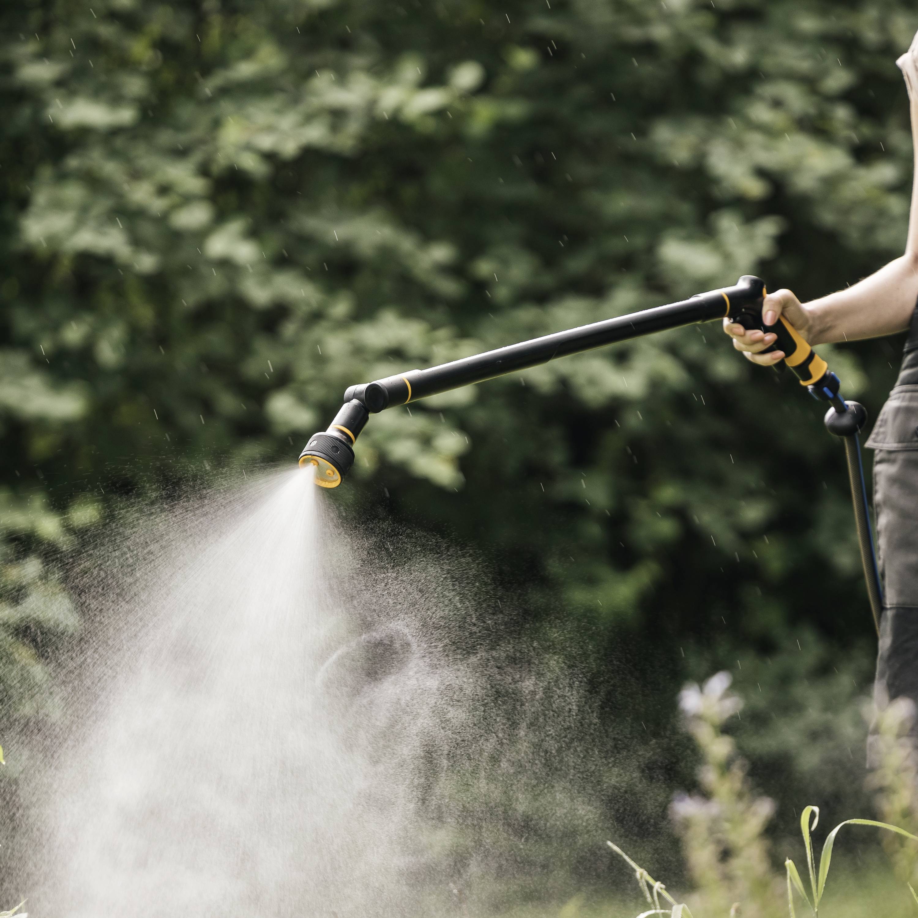Osoba podlewa rośliny w ogrodzie za pomocą węża ogrodowego. Strumień wody jest silny, a otoczenie jest bujnie zielone.