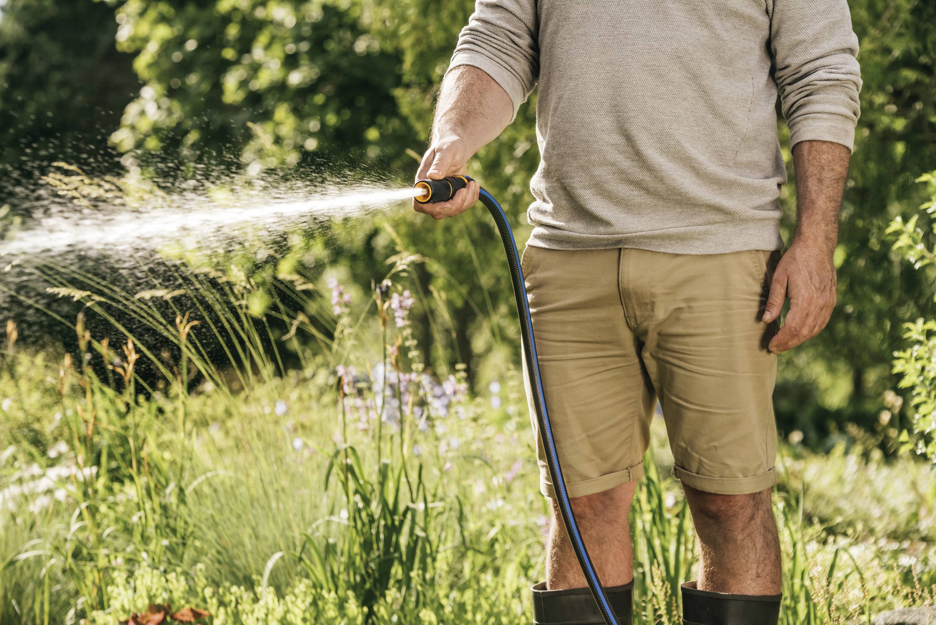 Mężczyzna podlewa kwitnący ogród za pomocą węża. Ubrany jest w krótkie spodenki i gumowce, otoczony zielenią roślin.