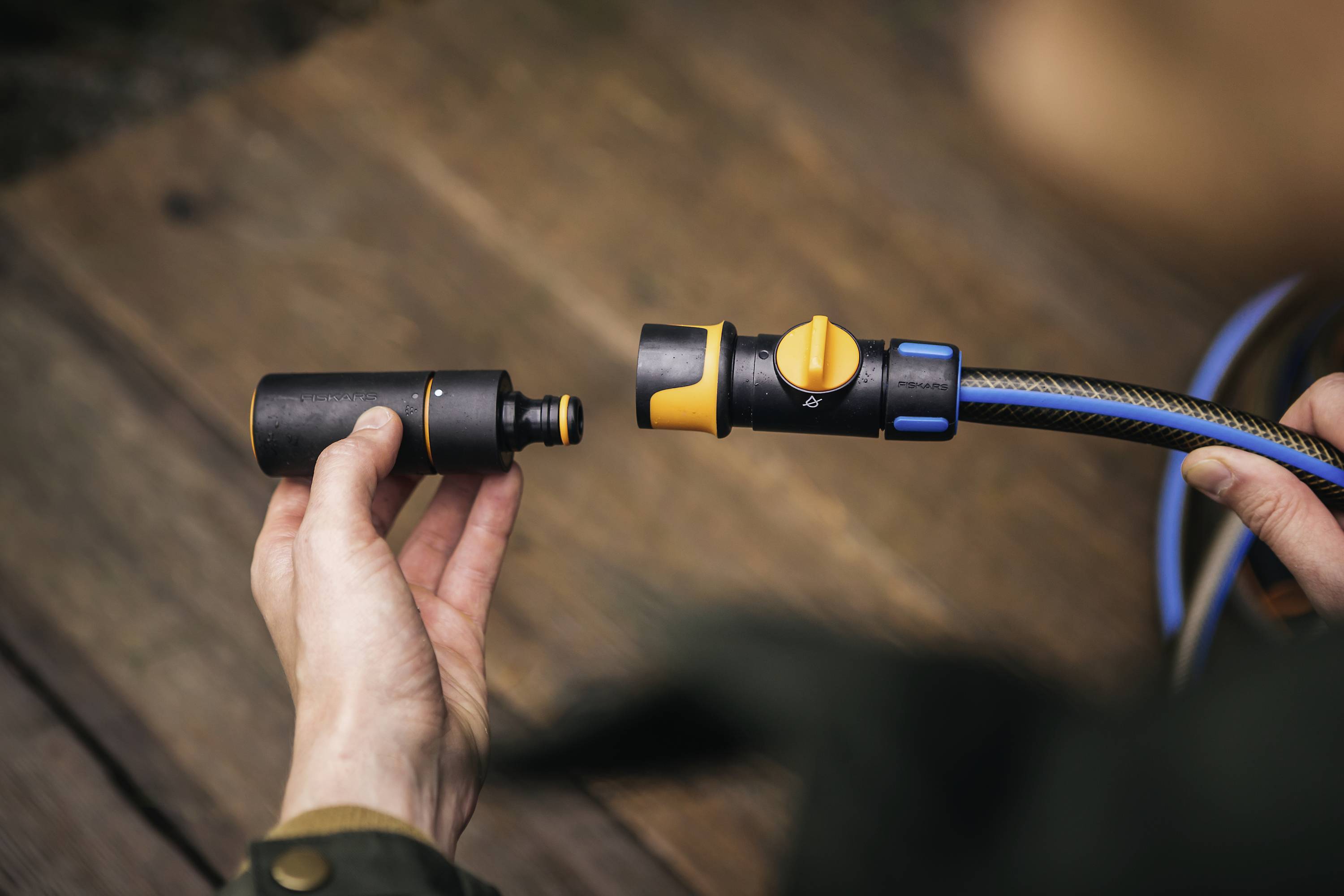 Osoba trzyma w ręku złączkę węża ogrodowego i podłącza ją do czarnego adaptera na drewnianej powierzchni.