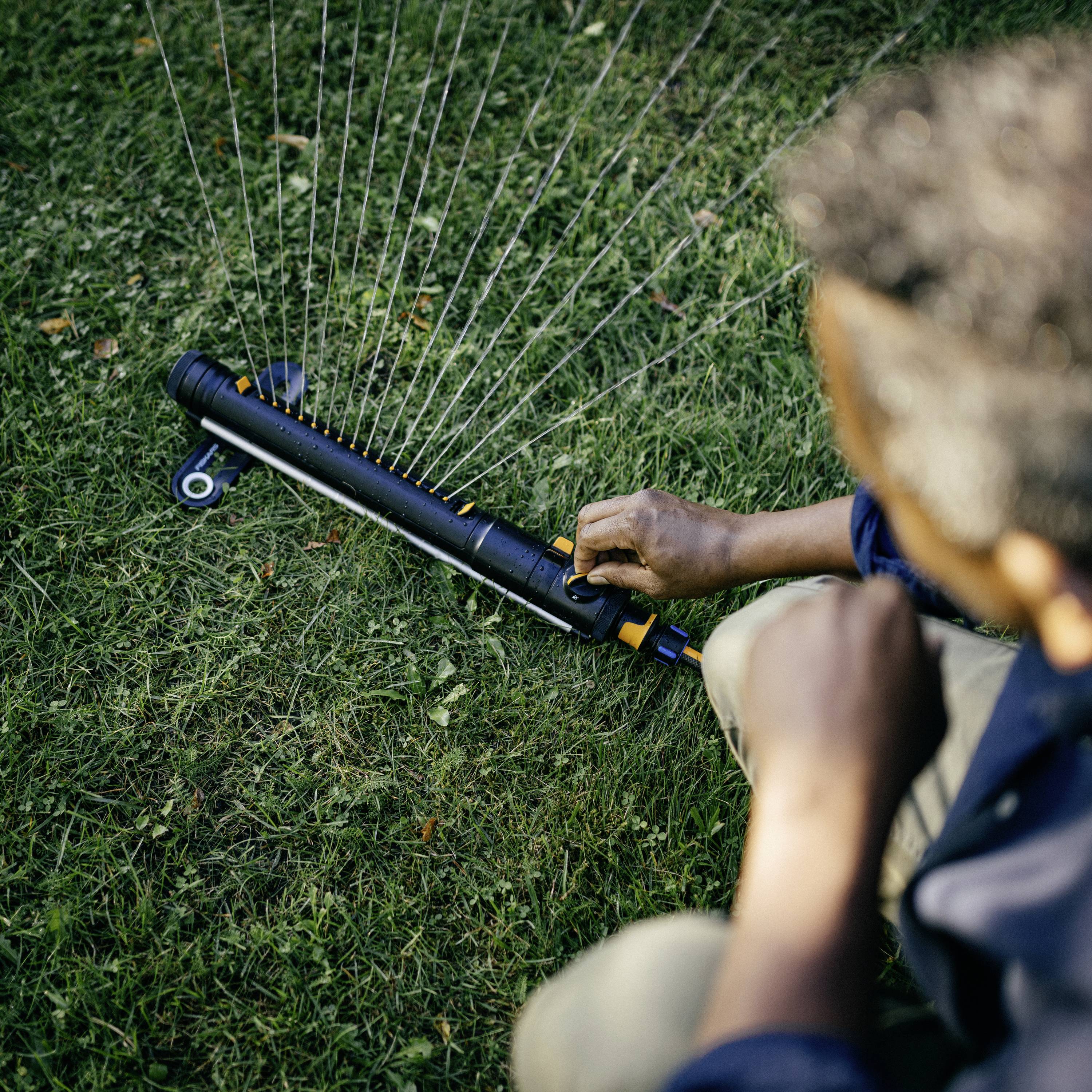 Osoba obsługuje zraszacz na zielonym trawniku. Zraszacz rozpyla wodę w wachlarzowatym wzorze.