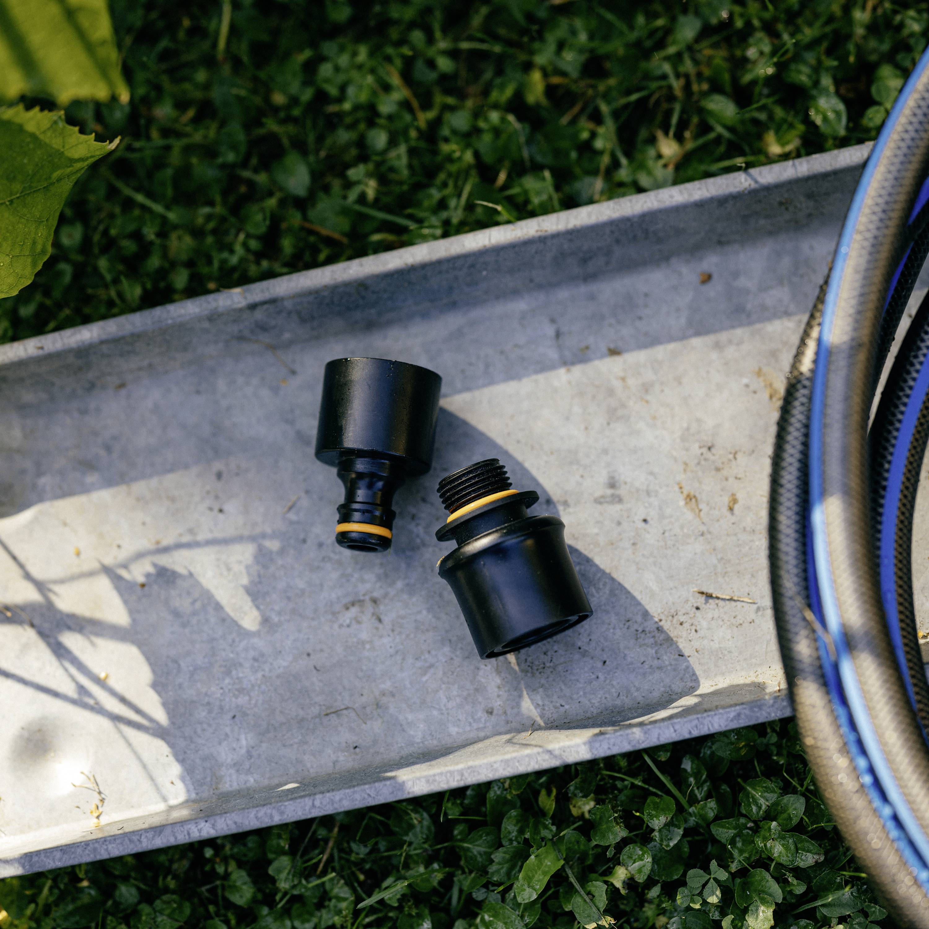 Złączka ogrodowa i adapter leżą na metalowej tacy na trawie. Po prawej stronie obrazka widać fragment niebieskiego węża.