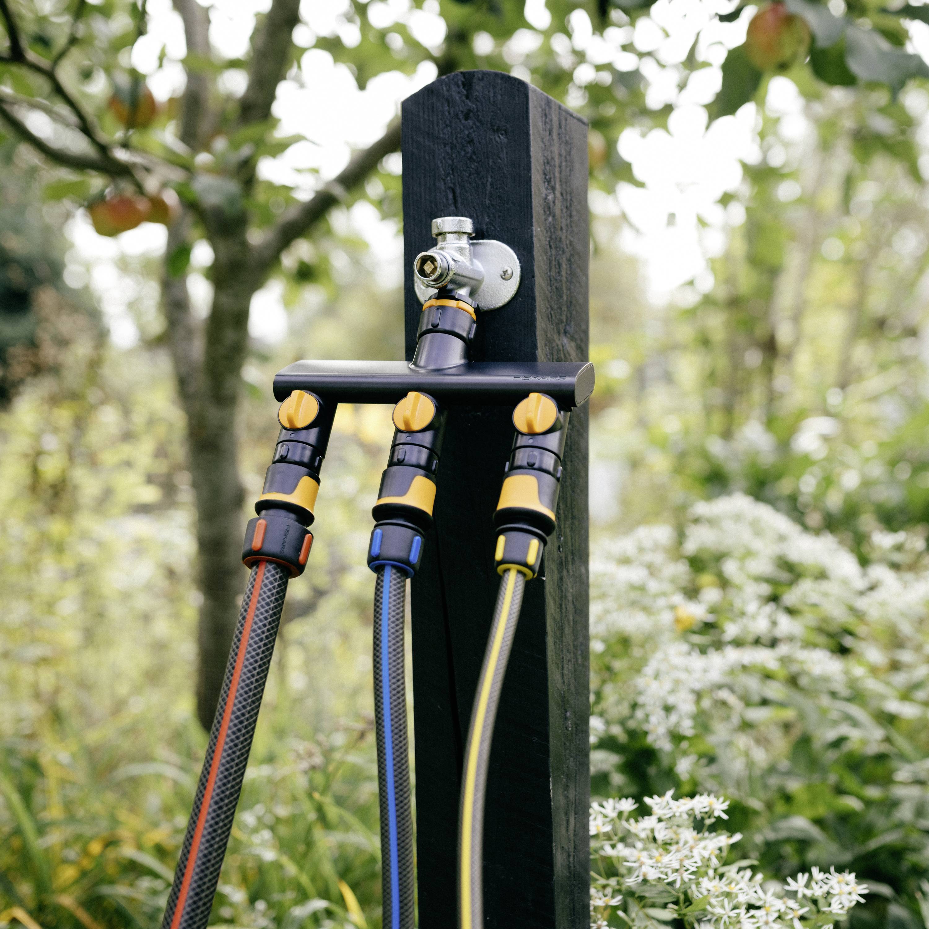 Trzy węże ogrodowe są przymocowane do rozdzielacza wody, który jest zamontowany na drewnianym słupie. W tle widać drzewa i rośliny.