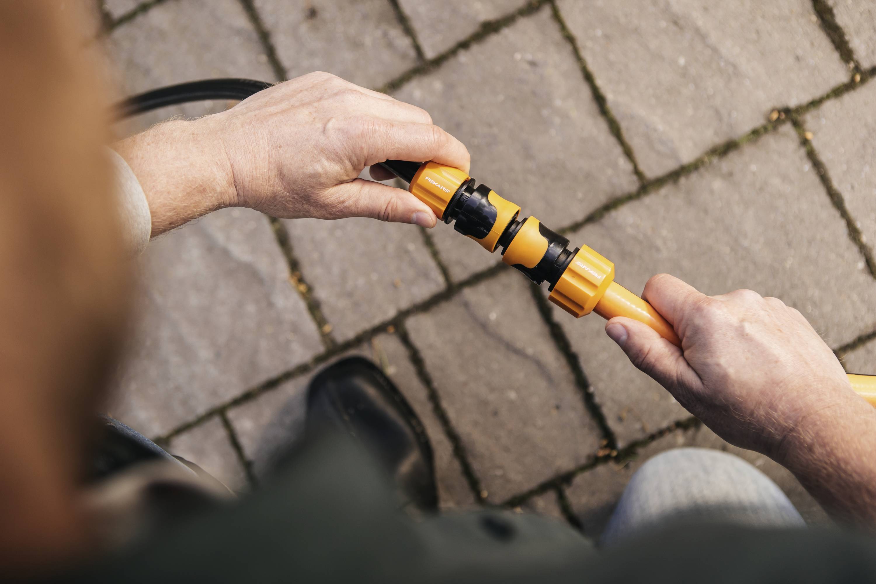 Osoba łączy węże ogrodowe żółtymi złączkami na wybrukowanej ścieżce. Fokus skierowany jest na dłonie i połączenie.