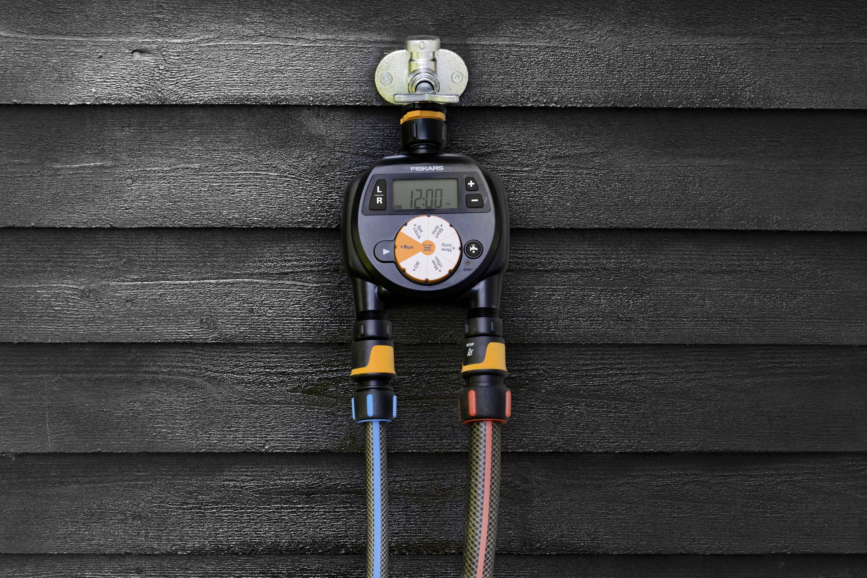 Licznik nawadniania z cyfrowym wyświetlaczem pokazuje '12:00'. Podłączone są dwa węże, jeden niebieski, drugi czerwony. Zamocowany na ścianie.