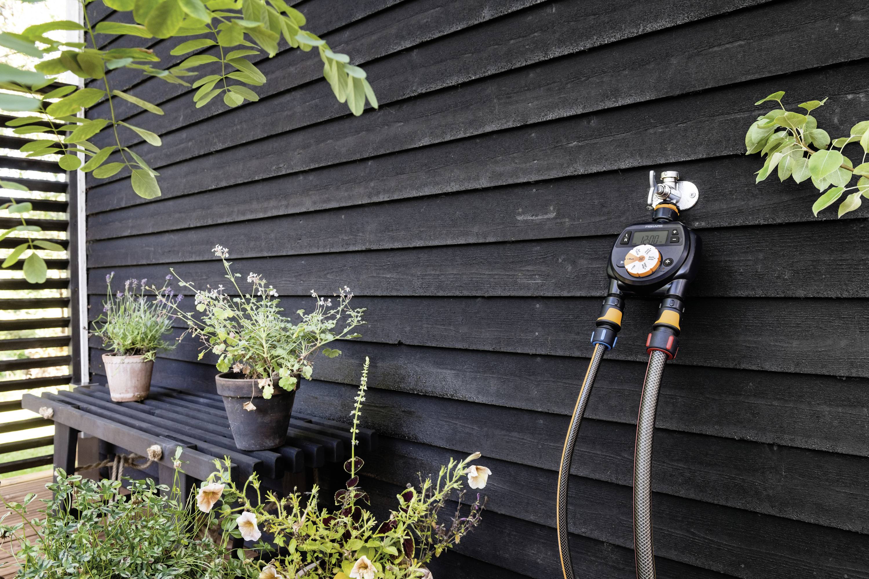 Czarna drewniana ściana z podłączonym systemem nawadniania obok roślin w doniczkach ustawionych na regale.