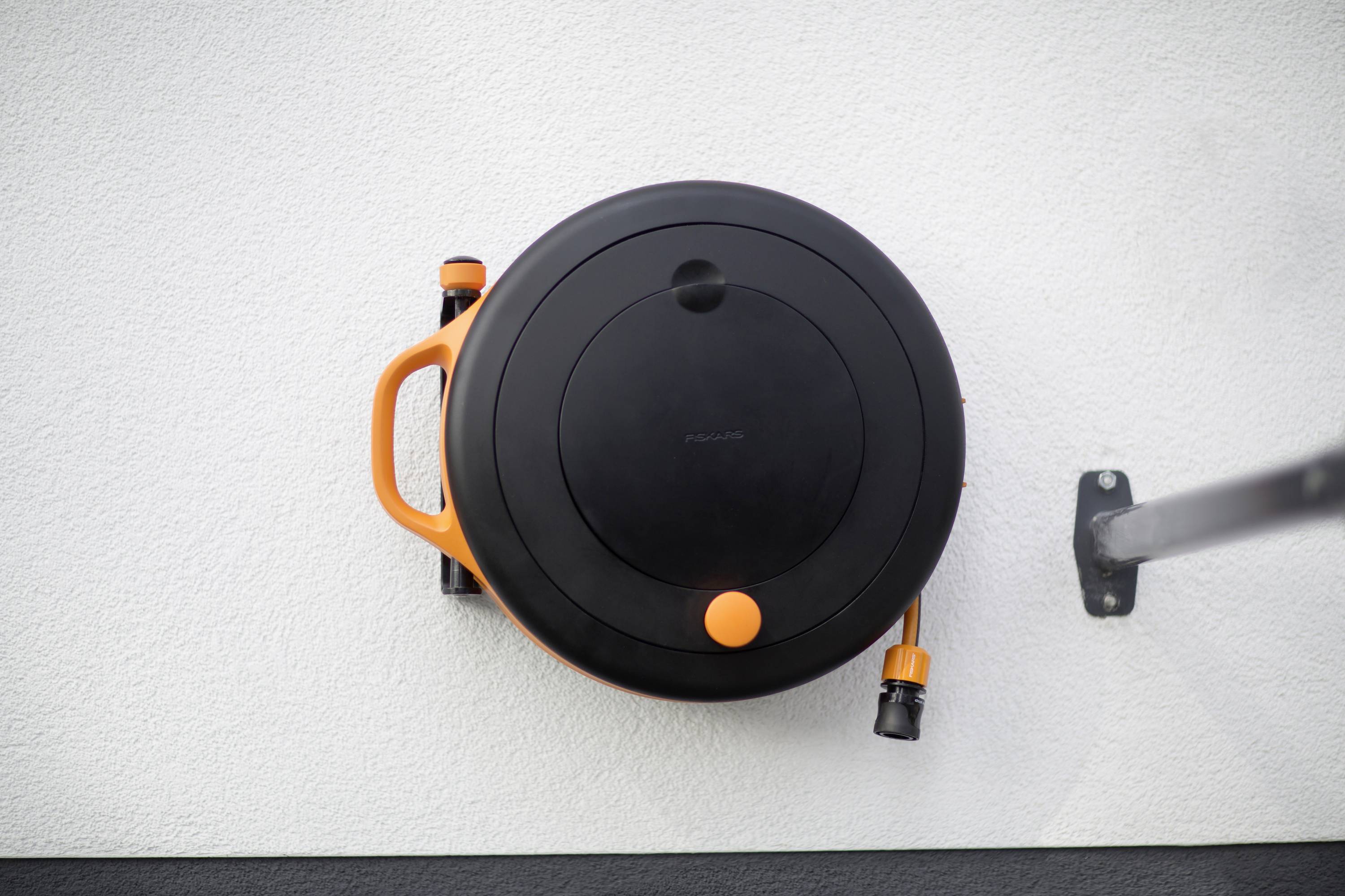 Okrągły, czarny wąż ogrodowy z pomarańczowymi akcentami jest zamontowany na białej ścianie.