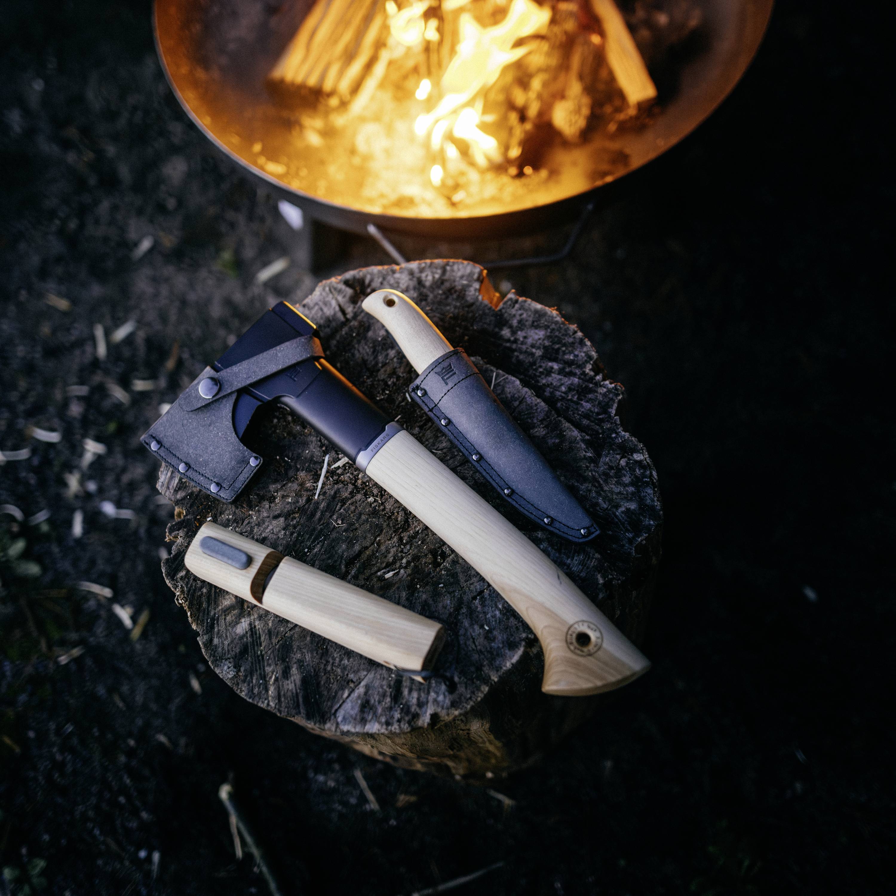 Siekiera i nóż z drewnianymi rękojeściami umieszczone na pniu drzewa przed ogniskiem obozowym. Narzędzia outdoorowe do kampingu i survivalu.