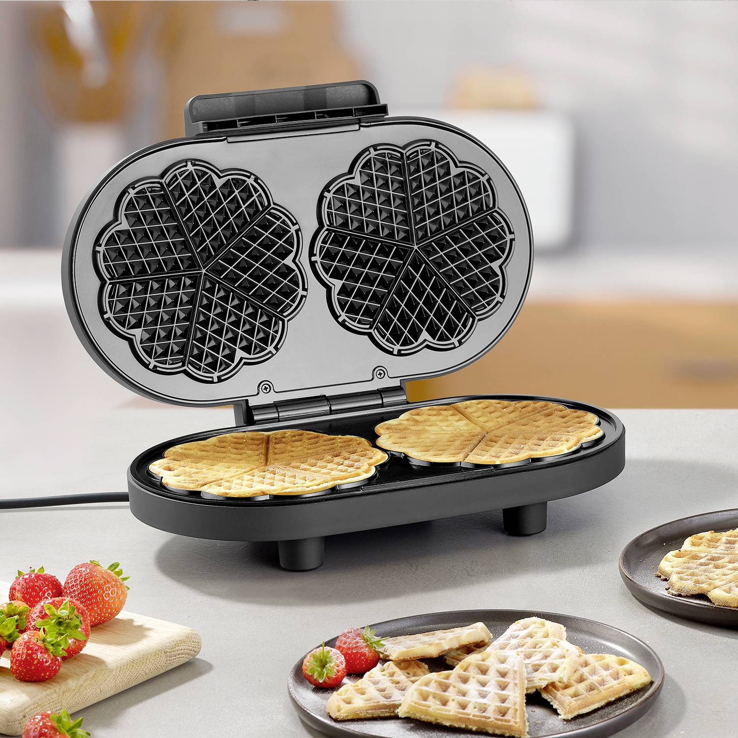 Żelazko do gofr z otwartą płytą, piecze dwie wafle w kształcie serc. Obok na talerzu leżą świeże gofry i truskawki.