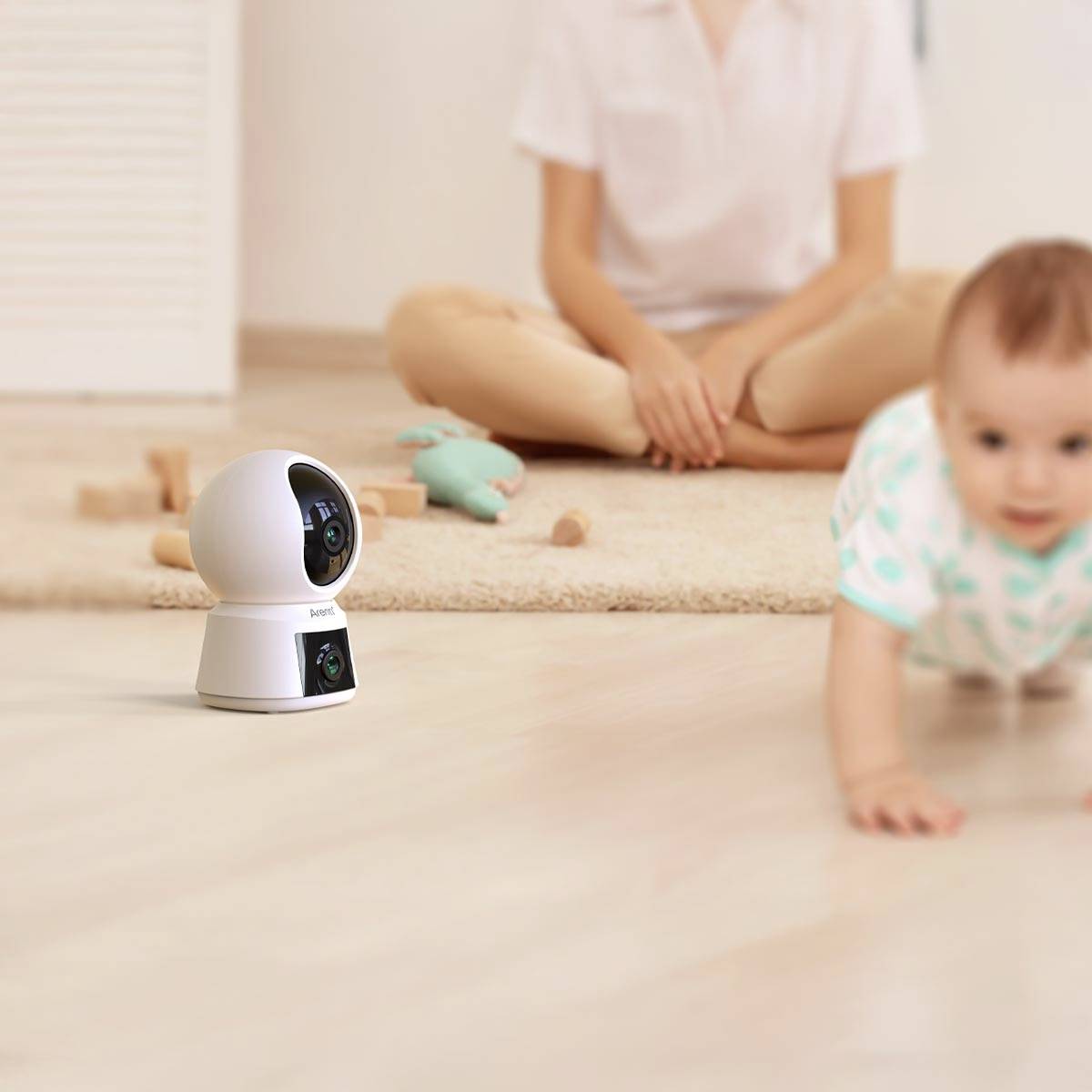 Niemowlę pełza po dywanie, podczas gdy kamera bezpieczeństwa jest wyraźnie widoczna na pierwszym planie. W tle siedzi jakaś osoba.