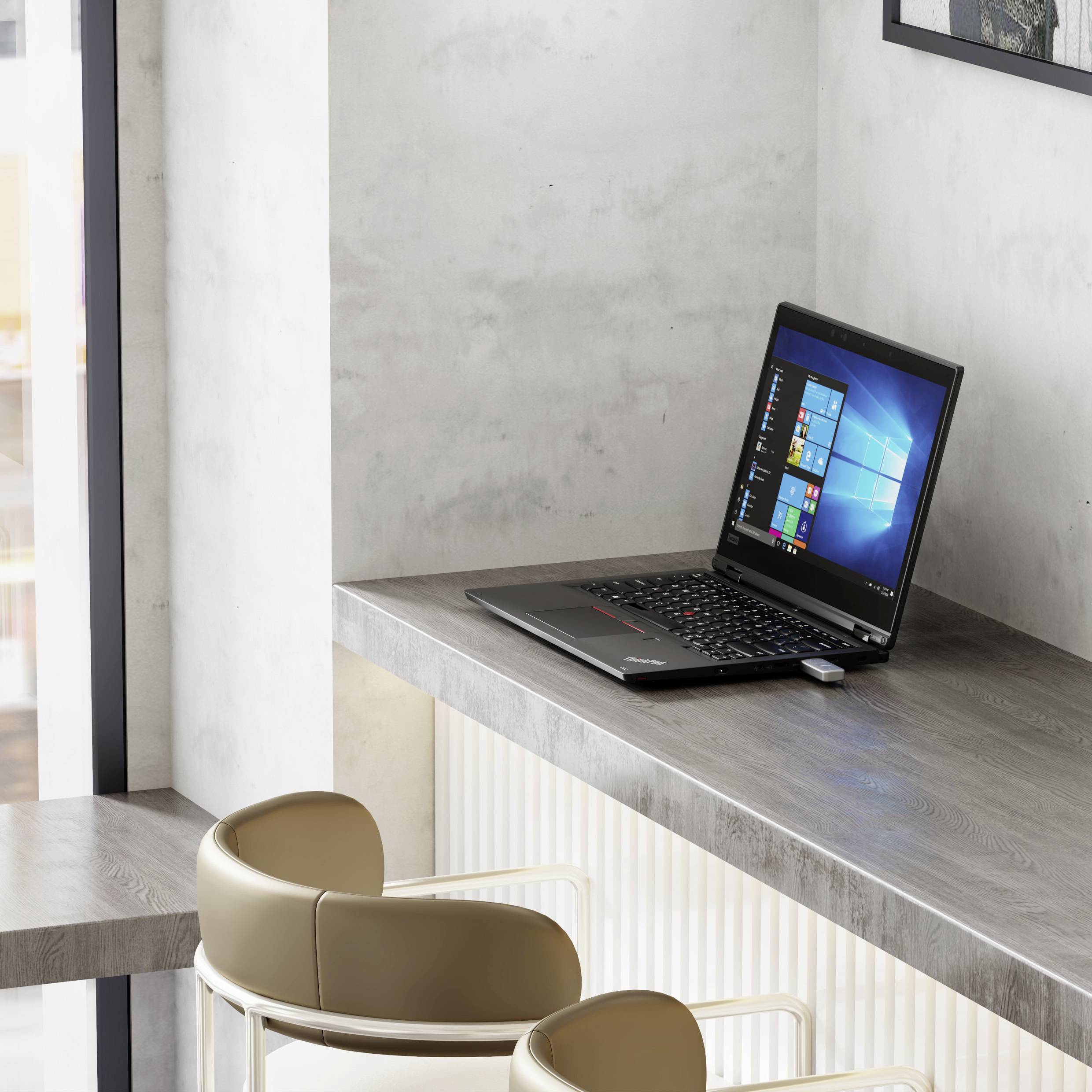 Laptop z ekranem Windows stoi na małym szarym biurku w pobliżu okna, obok jasnego krzesła w nowoczesnym, minimalistycznym pokoju.