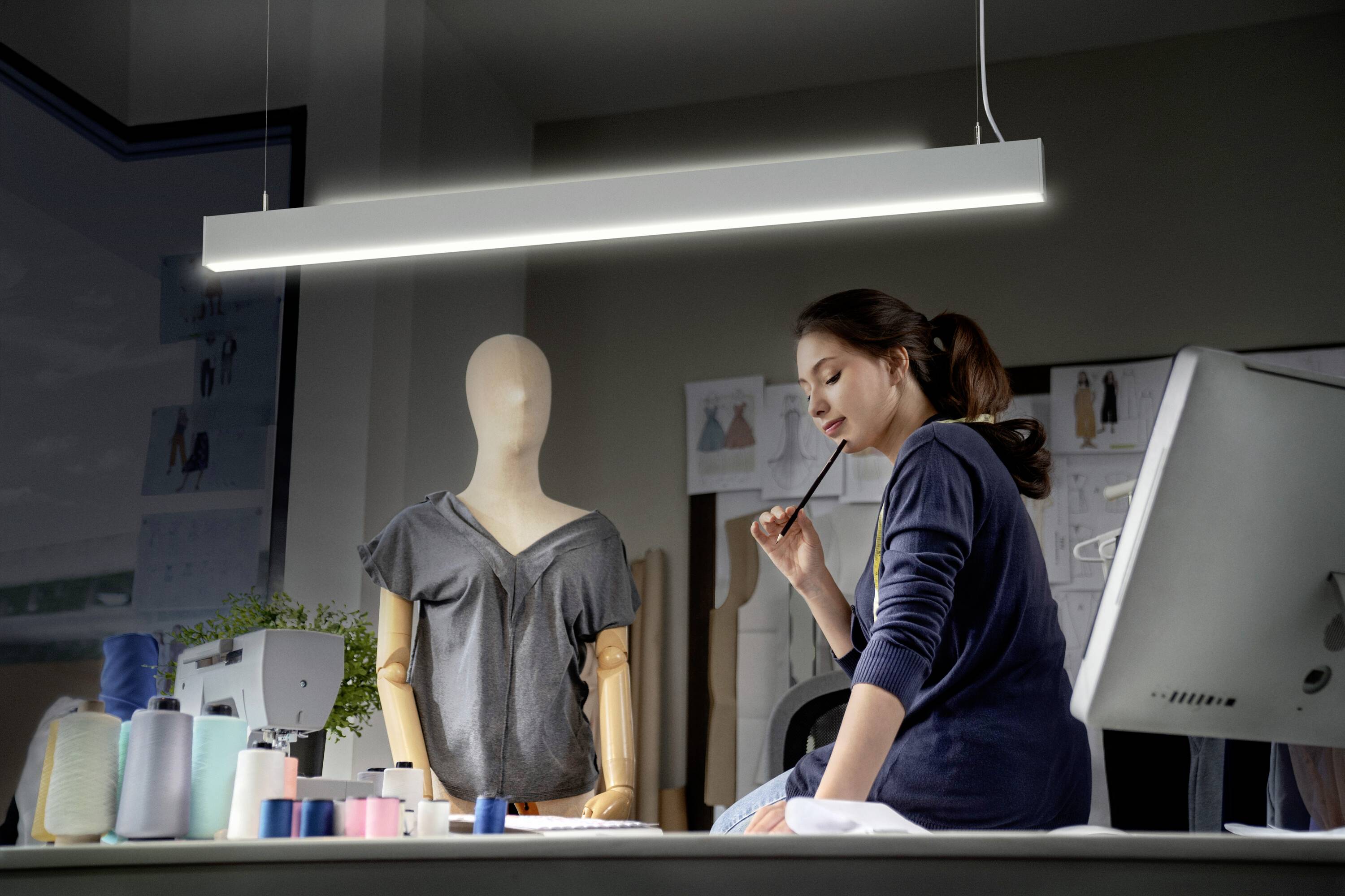 Kobieta siedzi przy biurku i zamyślona przygląda się manekinowi krawieckim z koszulką, otoczona materiałami do szycia.