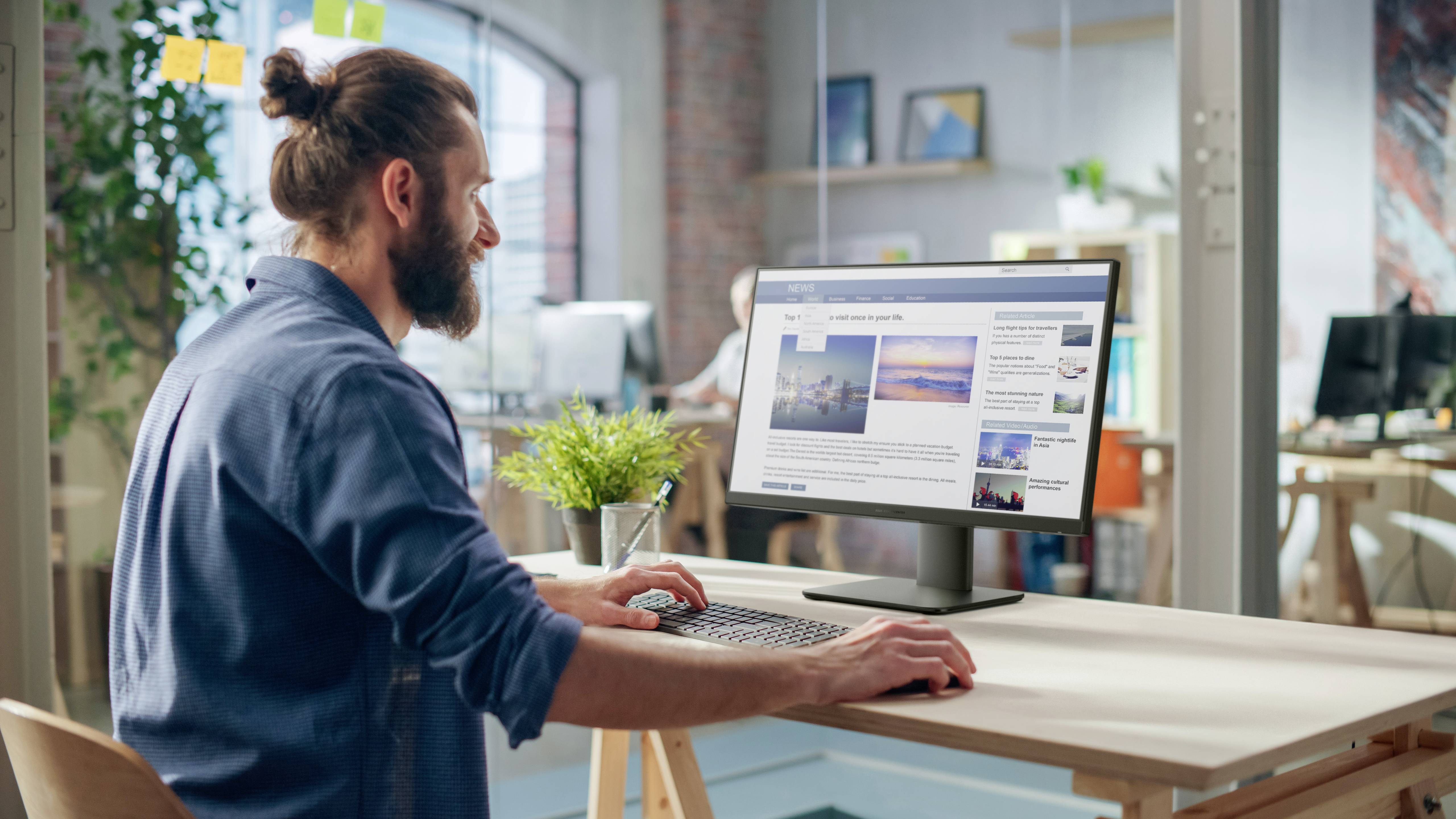 Osoba z brodą, siedząca przy biurku, patrzy na monitor komputera wyświetlający obrazy i tekst w nowoczesnym otoczeniu biurowym.