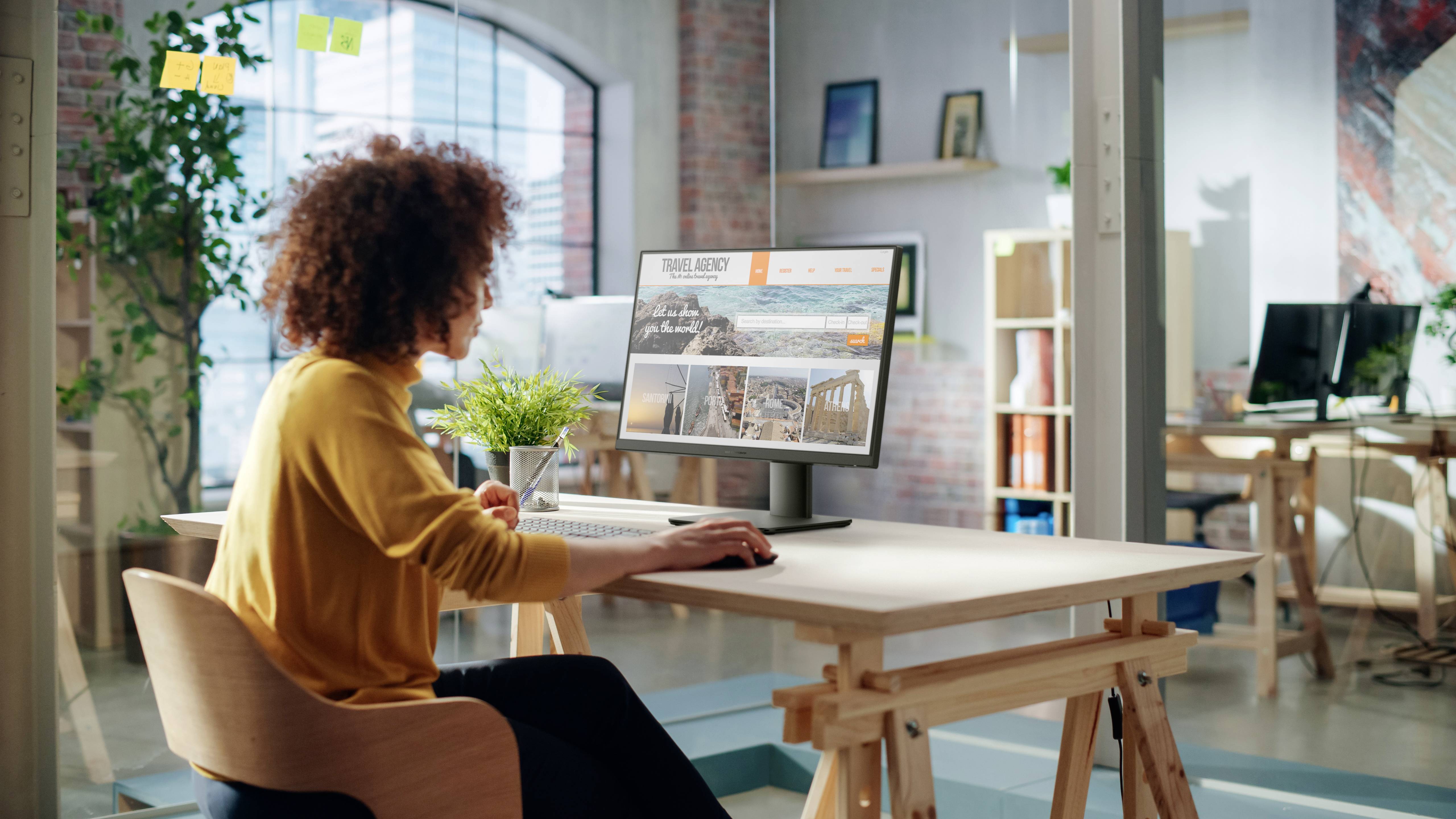 Osoba siedząca w jasnym biurze przy biurku patrzy na ekran komputera wyświetlający stronę internetową agencji podróży z różnymi zdjęciami destynacji.
