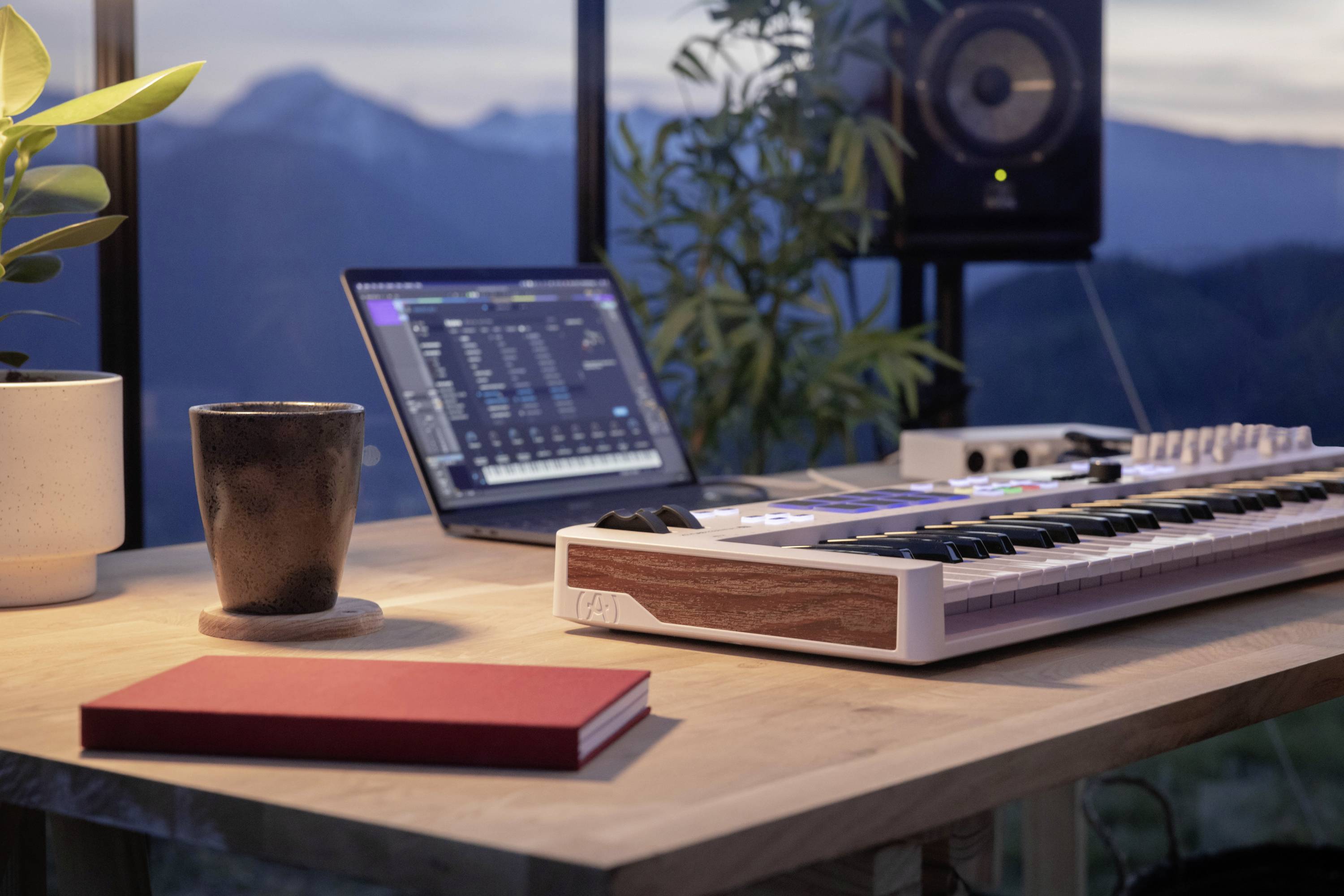 Laptop i klawiatura MIDI na biurku z widokiem na góry w tle; chłodne wieczorne światło i relaksująca atmosfera.