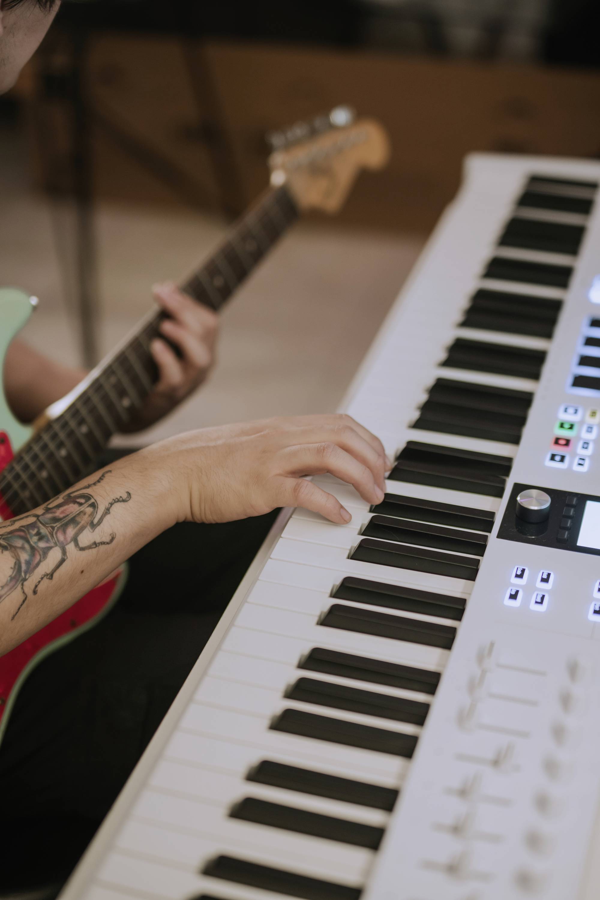 Osoba gra na gitarze elektrycznej z wytatuowanym przedramieniem i jednocześnie naciska klawisze białego keyboardu.