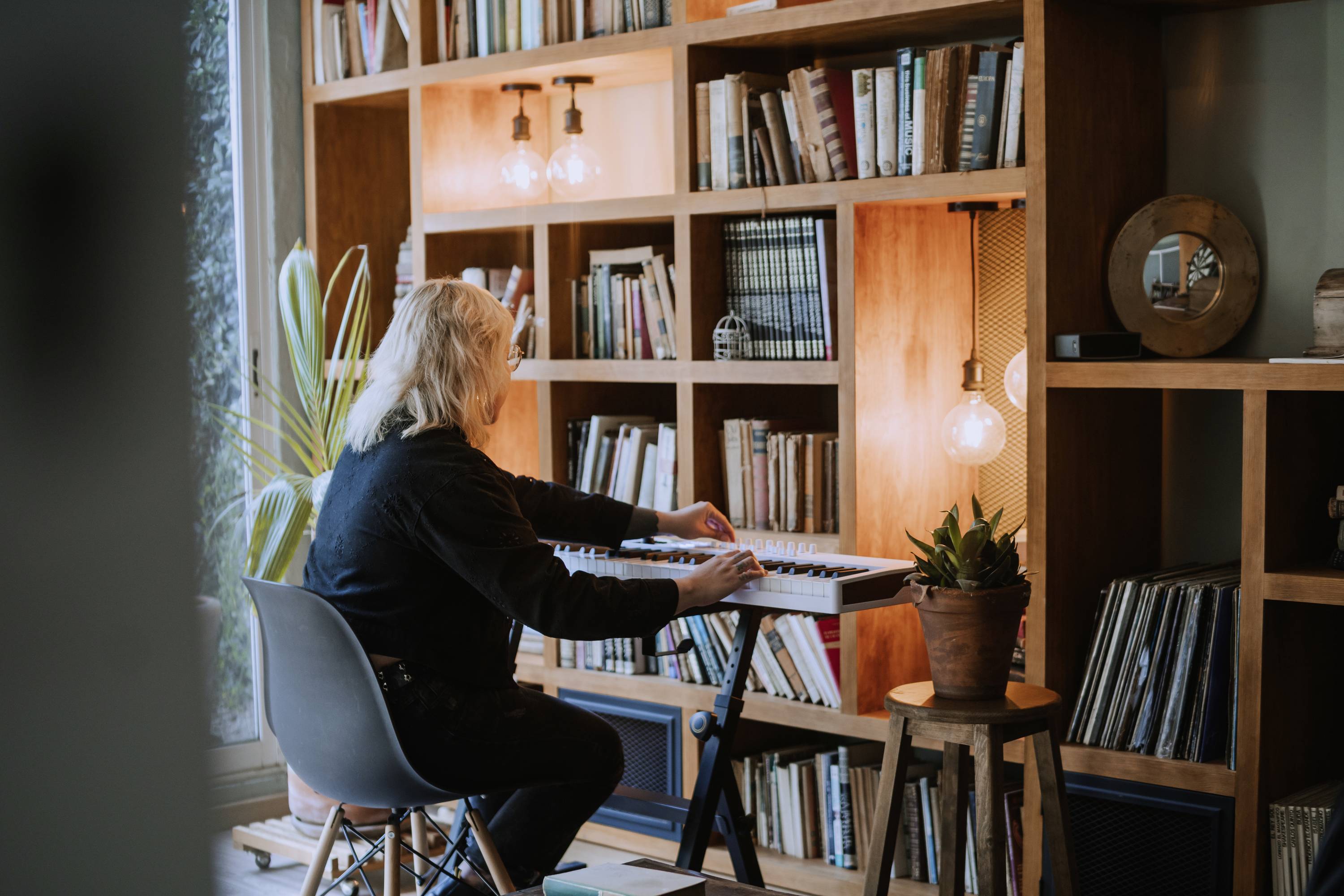 Osoba gra na keyboardzie w przytulnym pokoju z regałami na książki i roślinami. Ciepłe światło lamp oświetla scenę.
