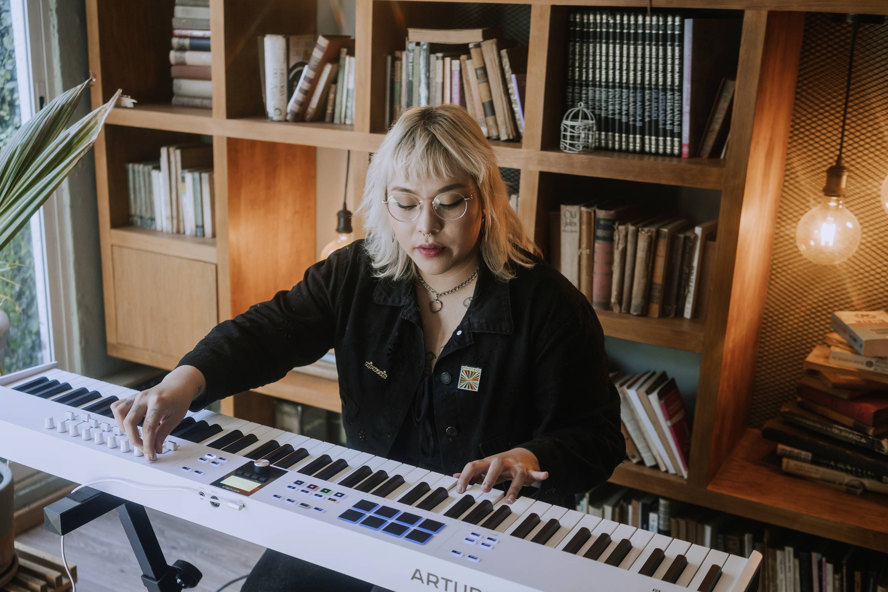 Osoba gra na białym keyboardzie w pomieszczeniu z półkami na książki w tle. Żarówka wisi na ścianie jako dekoracja.