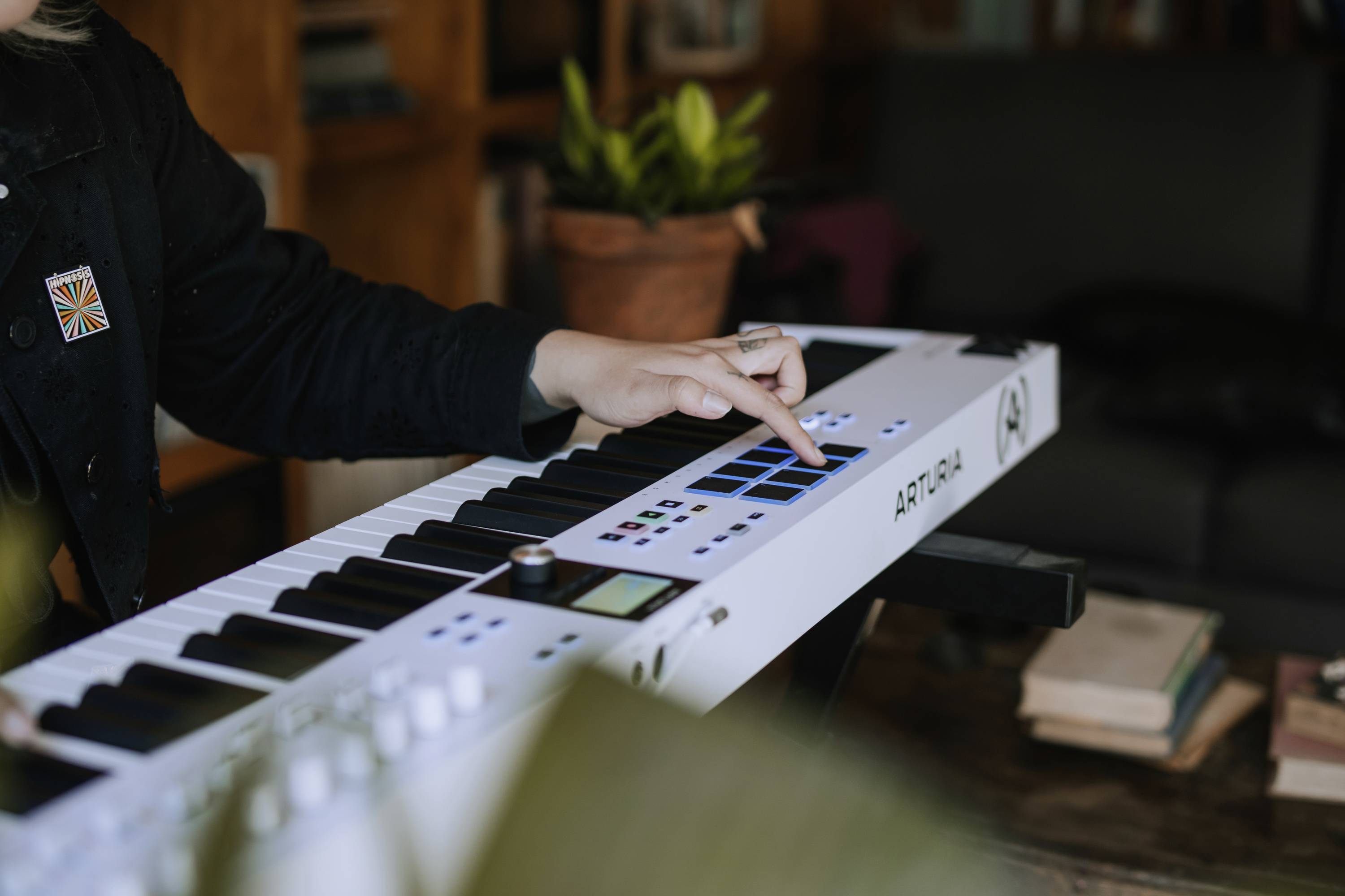 Osoba gra na białej klawiaturze MIDI w przytulnym pokoju. W tle widać książki i roślinę.