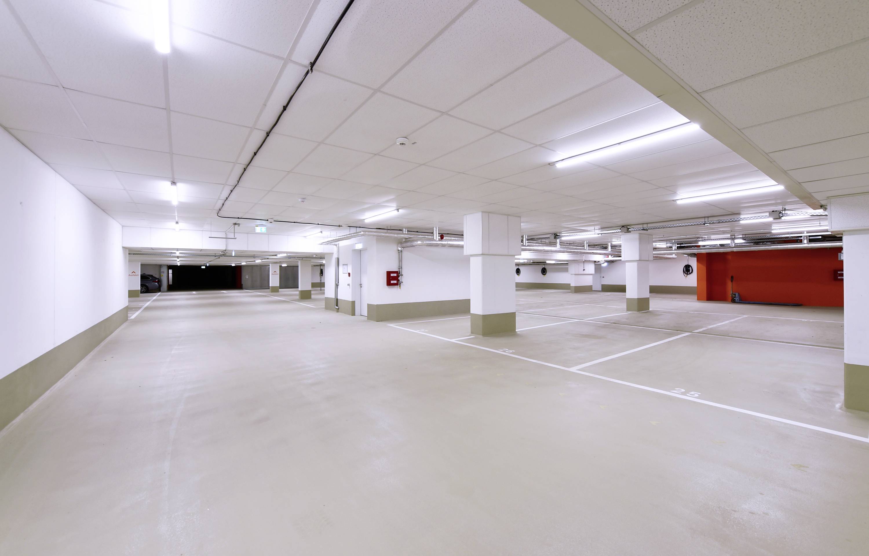 Pusty, dobrze oświetlony podziemny parking z białymi słupami, pustymi miejscami parkingowymi i czerwonym fragmentem ściany w tle.