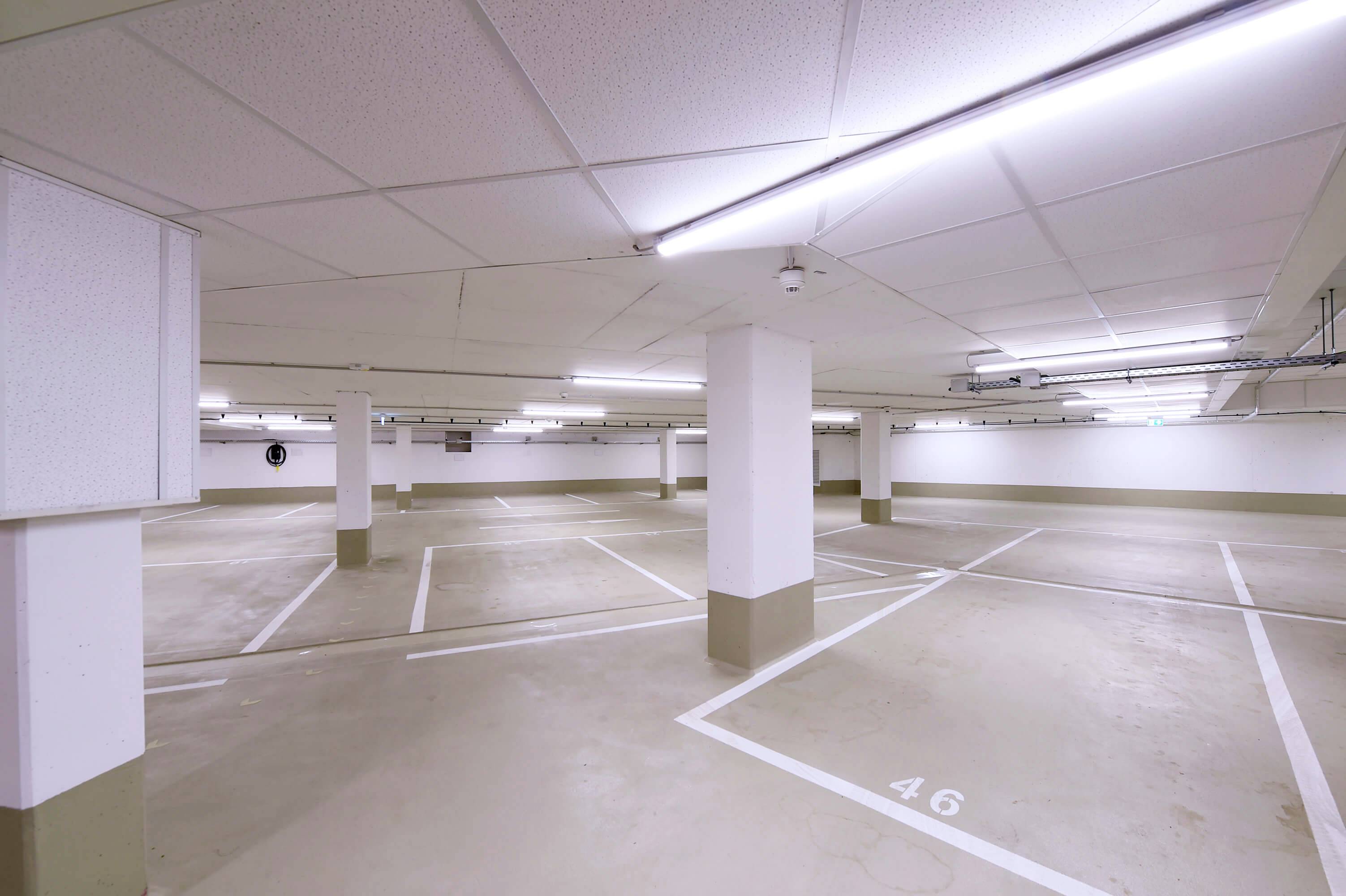 Puste miejsce parkingowe w garażu podziemnym z białym sufitem, oznaczeniami strzałek na podłodze i ponumerowanymi miejscami parkingowymi.