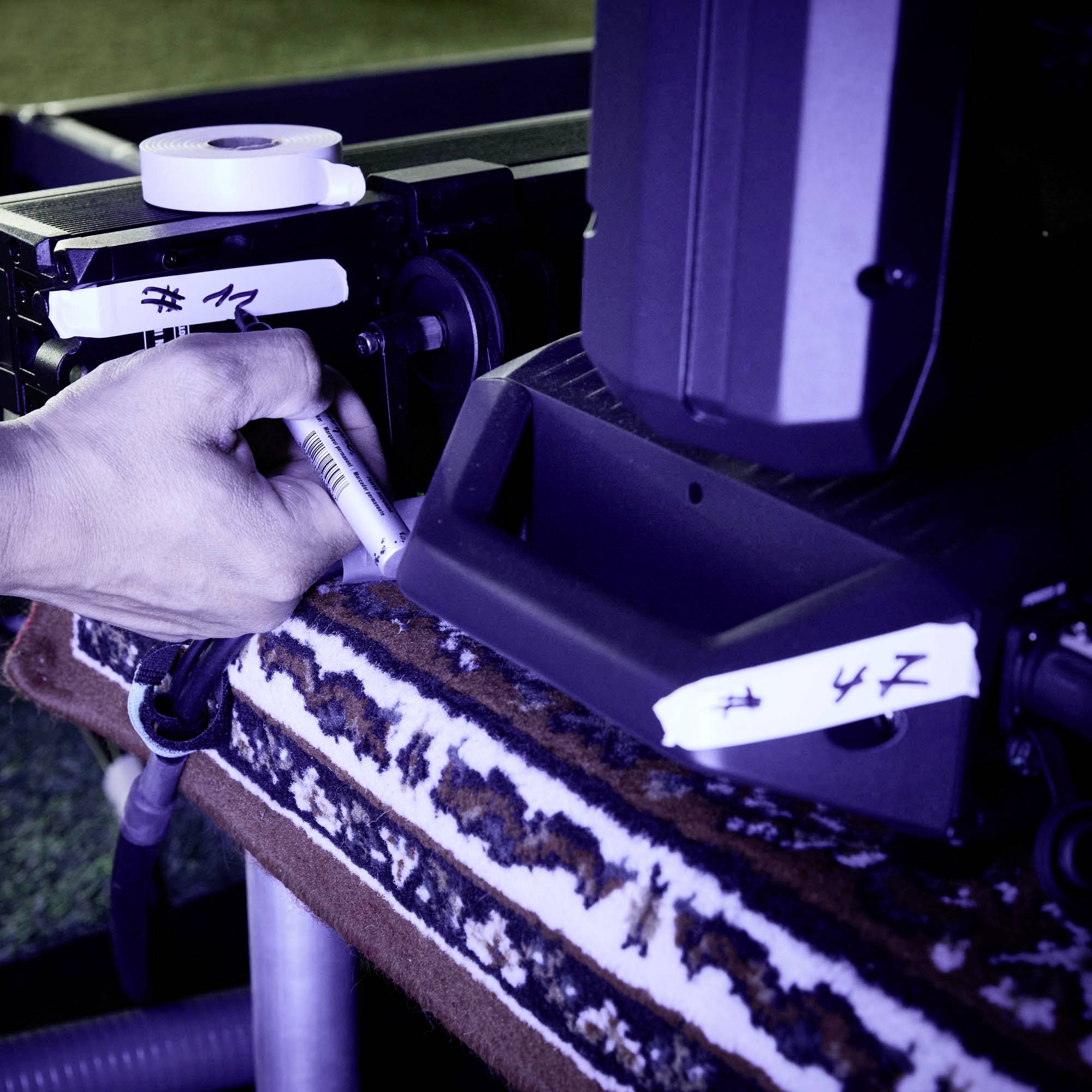 Ręka oznacza kamerę filmową białą naklejką. Scena ma niebieskie oświetlenie i pokazuje profesjonalny sprzęt filmowy.