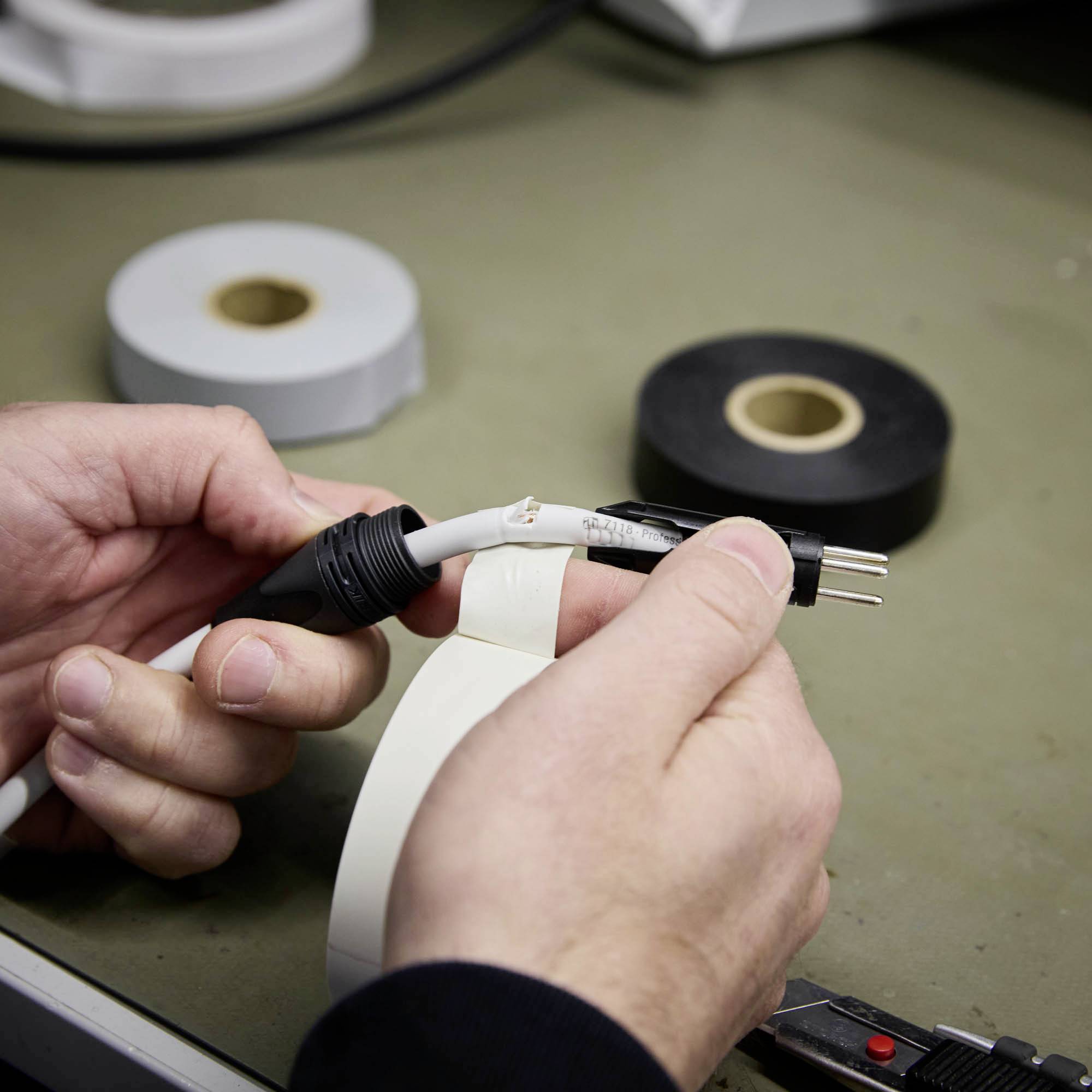 Osoba naprawia kabel elektryczny taśmą izolacyjną. W tle widoczne są dwie rolki taśmy klejącej, jedna biała i jedna czarna.