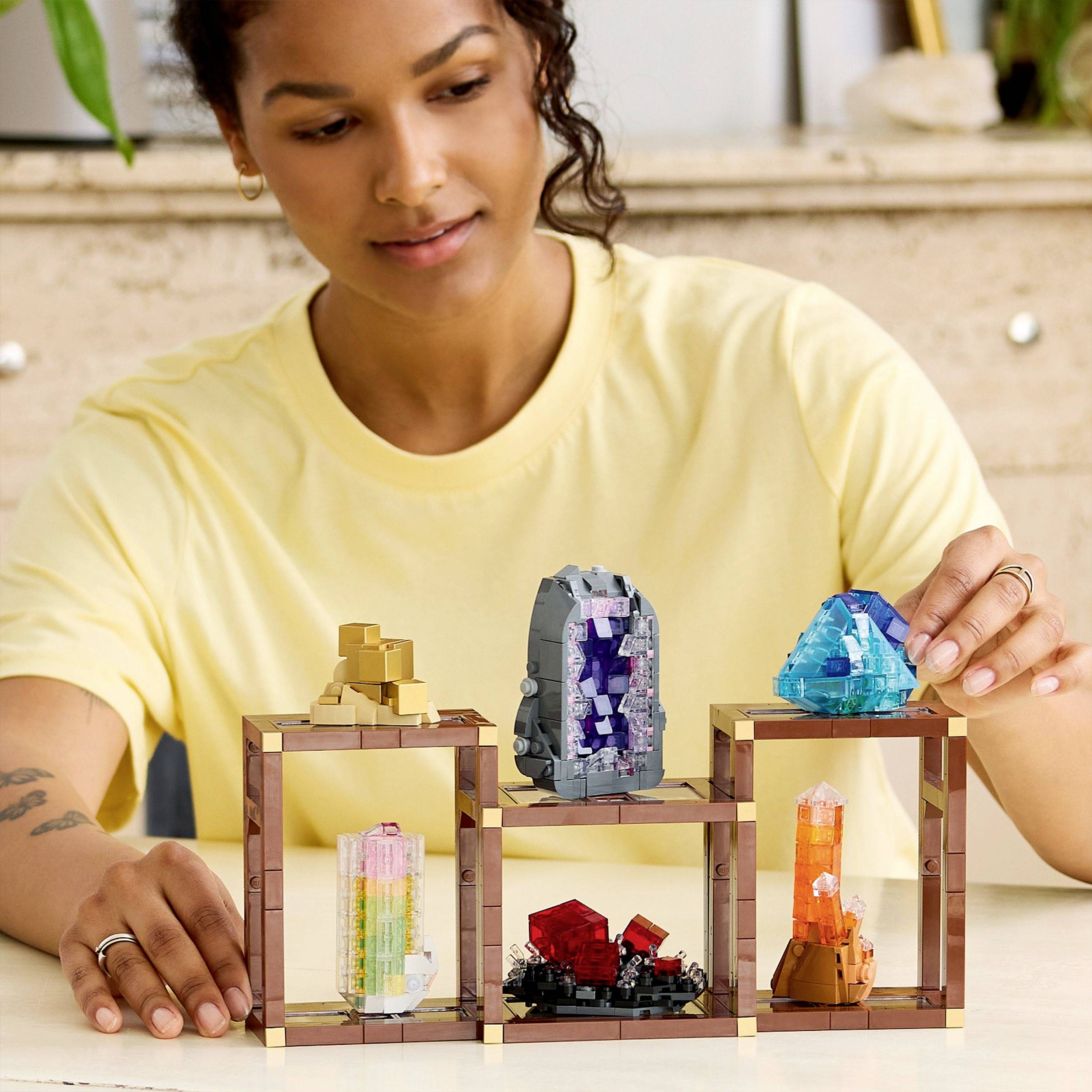 Osoba układa kolorowe, przypominające kryształy struktury na drewnianej półce, prezentując różnorodne geometryczne dzieła sztuki w pomieszczeniu.