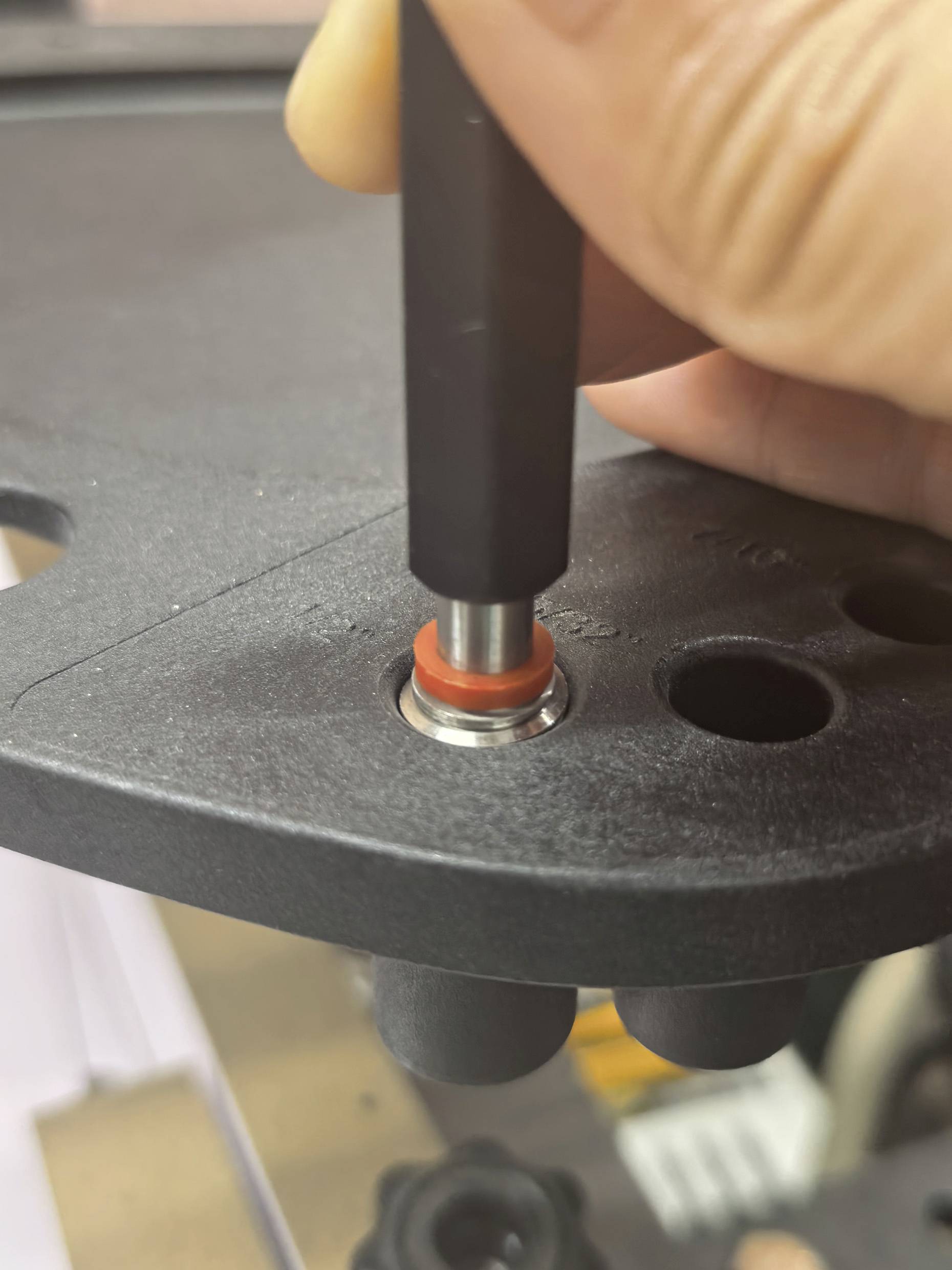Dłoń naciska na czarną plastikową powierzchnię za pomocą narzędzia z metalowym, spiczastym końcem, wyrównując je z okrągłą metalowo-gumową mocowaniem.