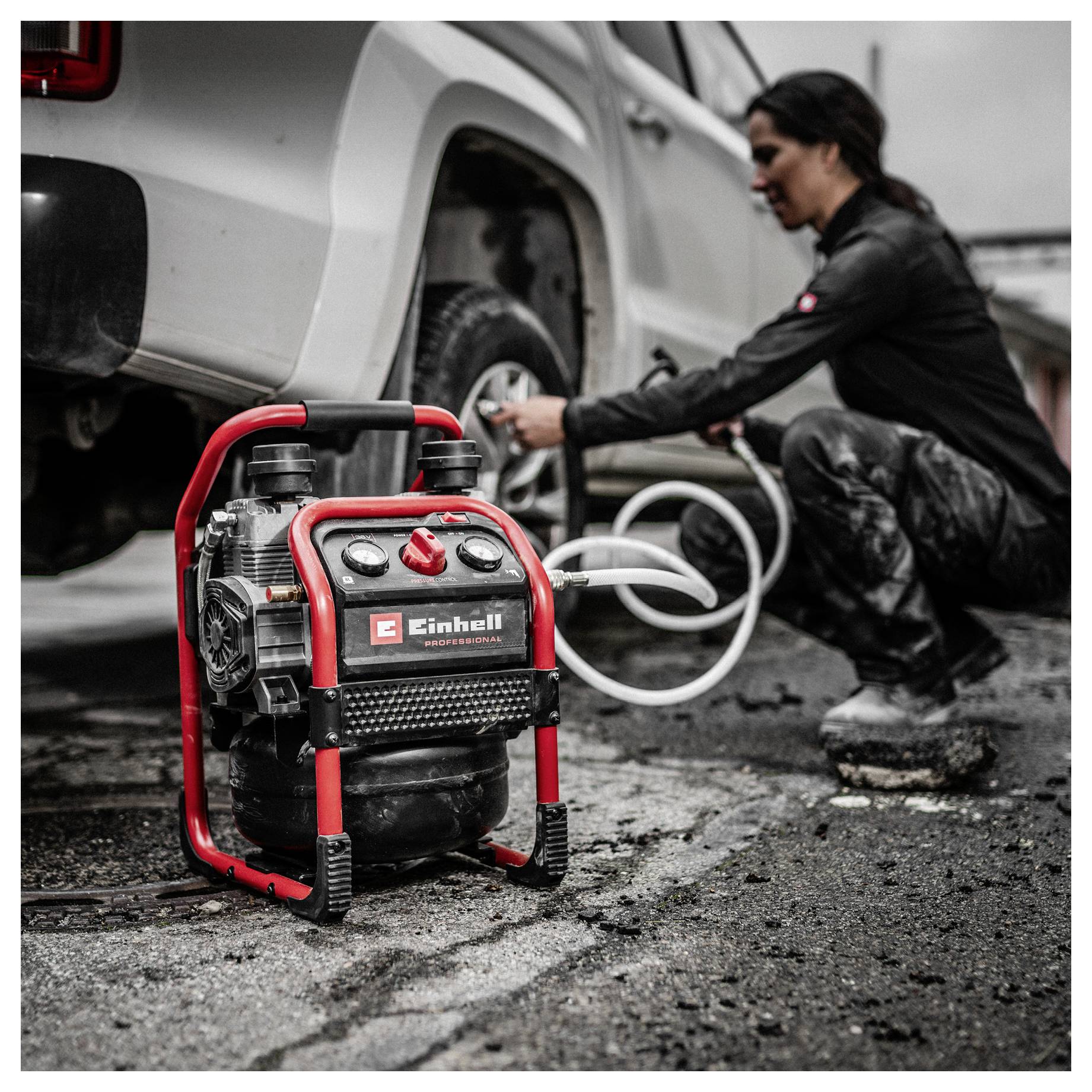 Przenośna pompa powietrza na pierwszym planie, podczas gdy kobieta używa jej do napompowania opony obok białej ciężarówki pikup na mokrym asfalcie.